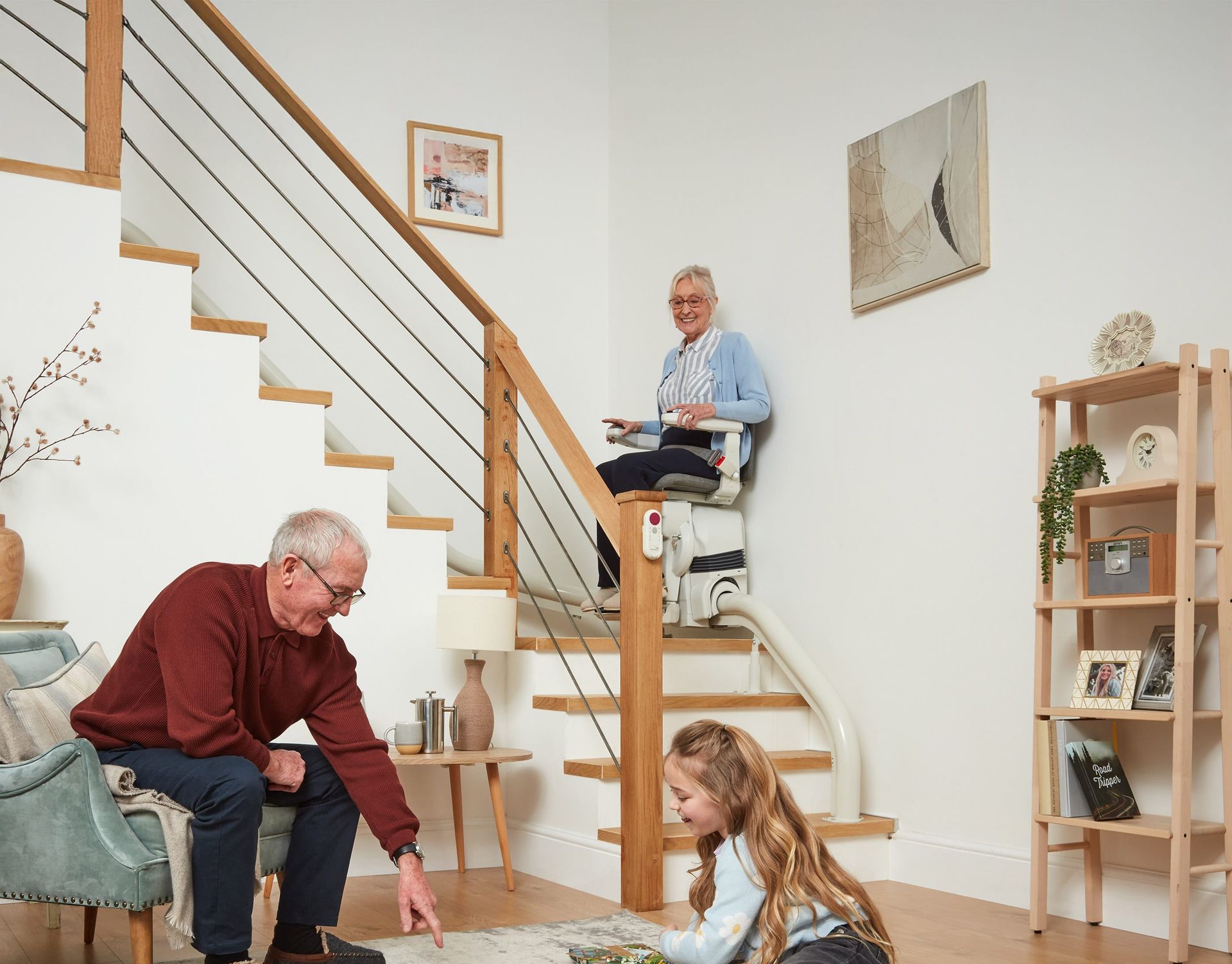 An older person on a stairlift, a man and child at the bottom of stairs, in a bright home setting.