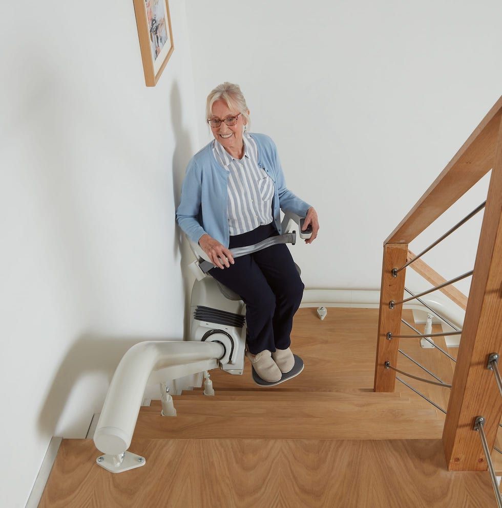 Person seated on a stairlift at the top of a wooden staircase, holding the armrest.