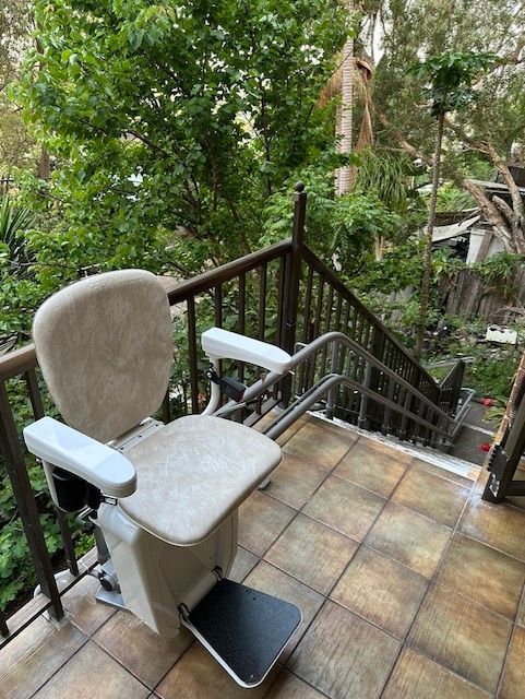 Stair lift installed on outdoor staircase. Beige chair with armrests, mounted on wall alongside steps.