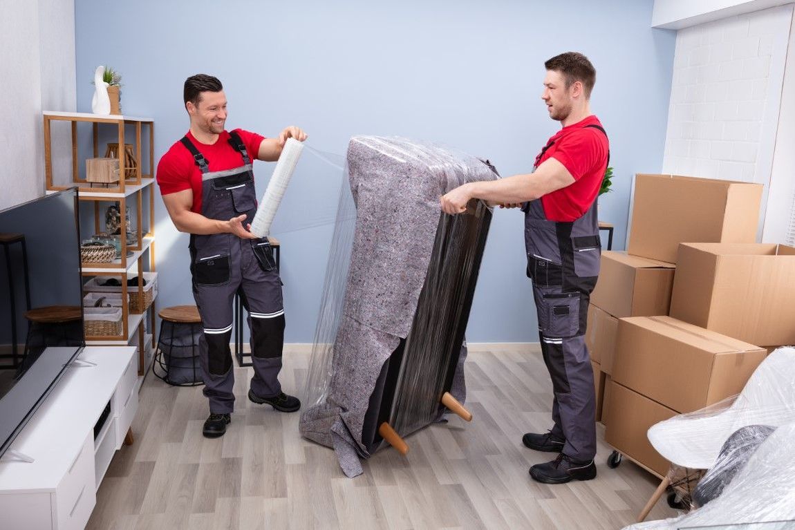 Two movers wrapping a couch in a living room, wearing red shirts and grey overalls.