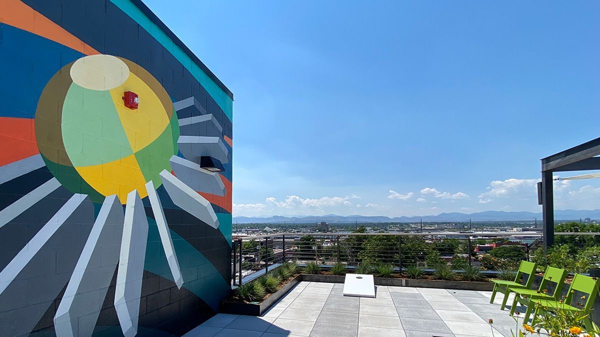 Rooftop mural with colorful sunburst design, overlooking a cityscape and mountains; lime green chairs.