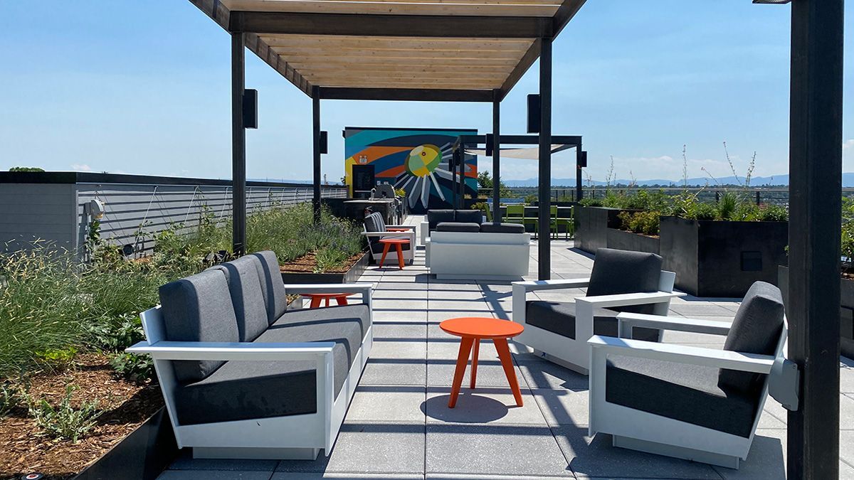 Rooftop patio with white couches, orange table, shaded pergola, and mural on a clear, sunny day.