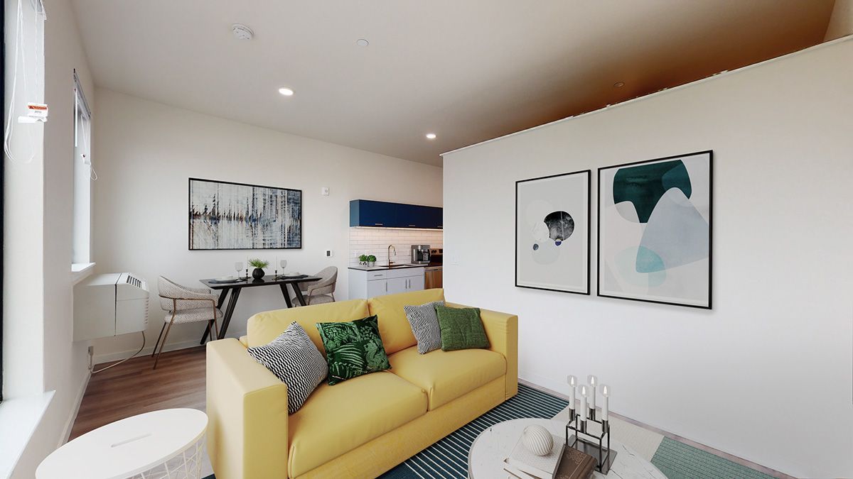 Living room with yellow couch, art prints, rug, and small dining area in a white-walled space.