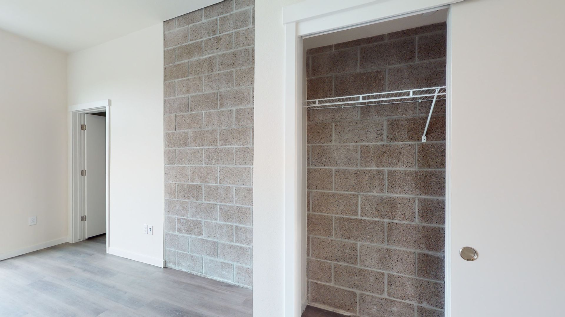 Interior room with exposed brick wall, closet, and a closed door in the background. White trim and gray flooring.