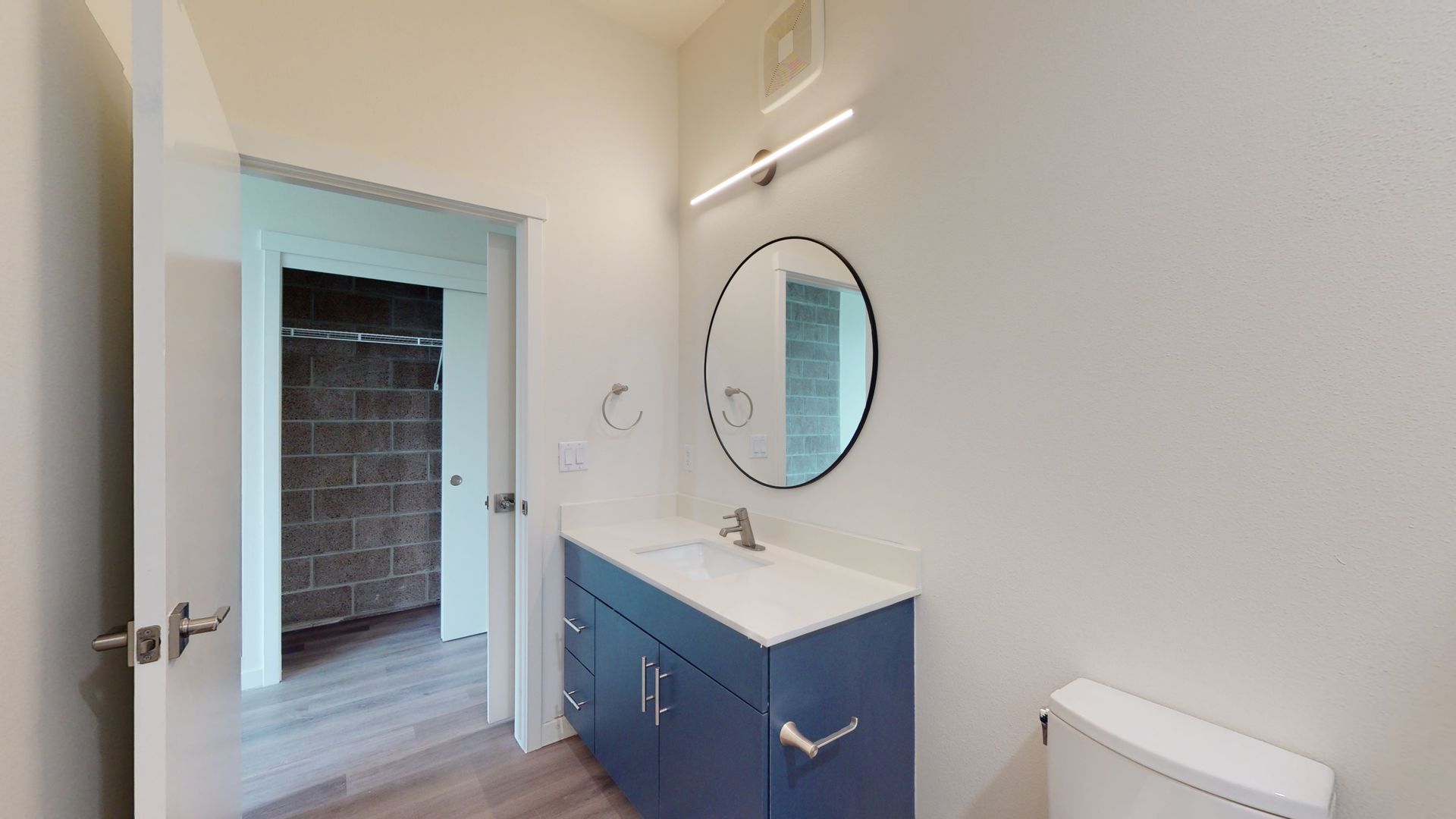 Bathroom with blue vanity, round mirror, and doorway to a dark room with brick wall.