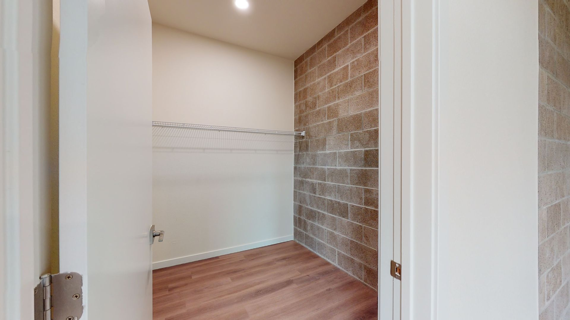 Empty closet with brick accent wall, white walls, wire shelf, wooden floor.