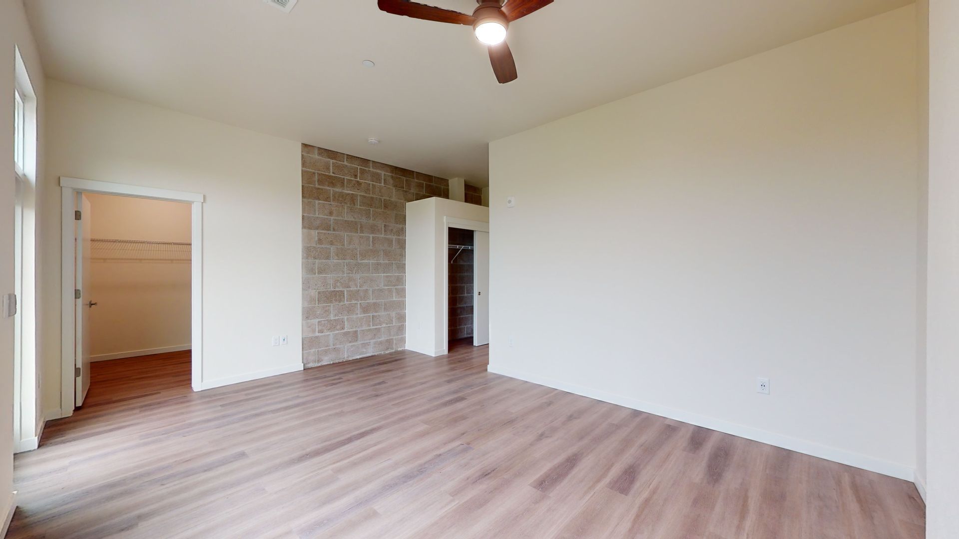 Empty room with wood floors, walk-in closets, and exposed brick wall. A ceiling fan is visible.