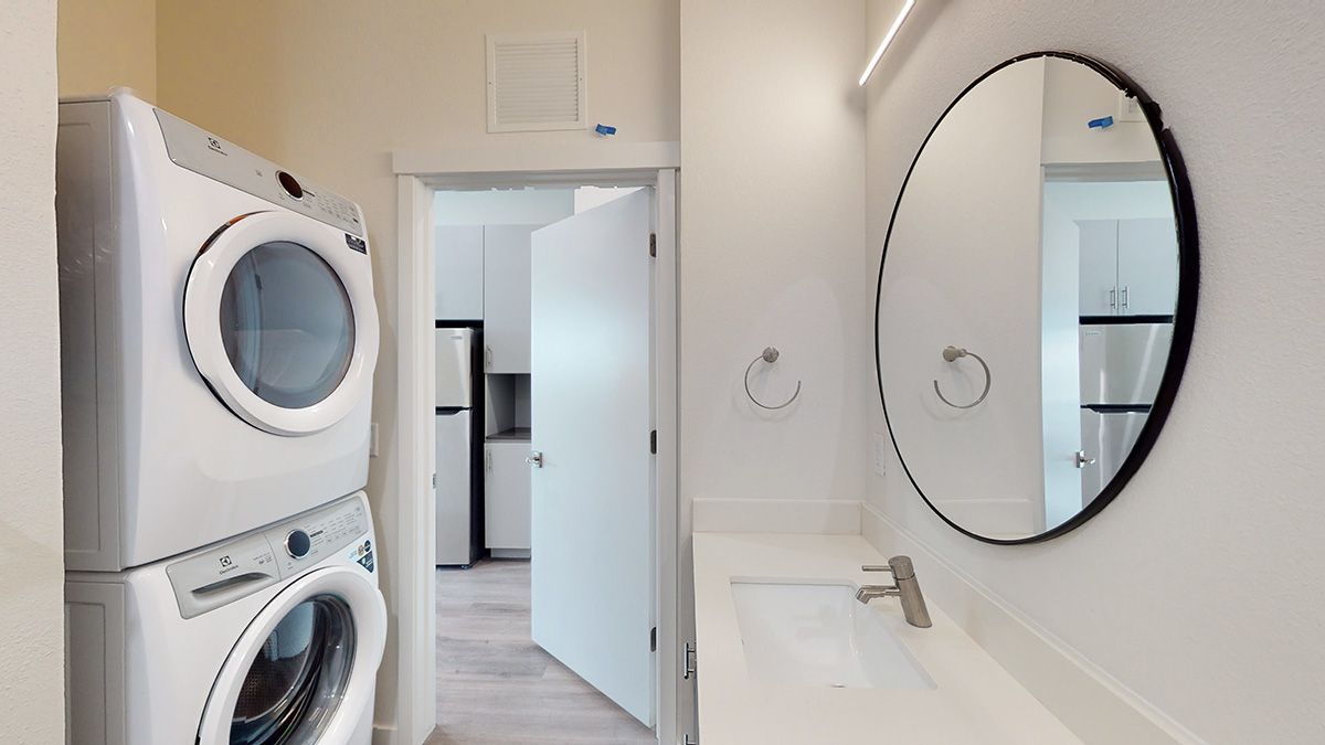 Laundry area with stacked washer and dryer, adjacent to a bathroom with a round mirror.