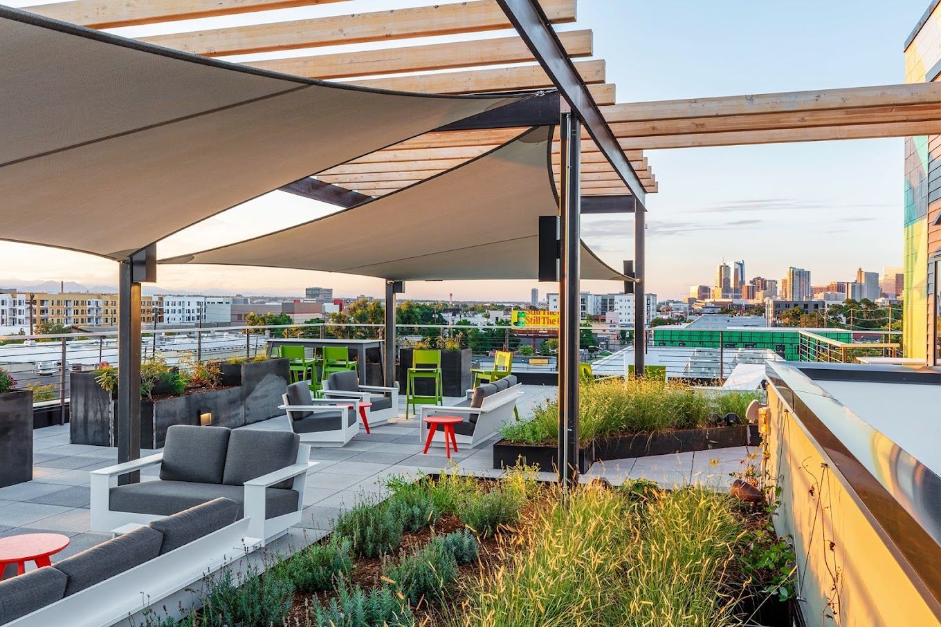 Rooftop lounge with shade sails, seating, and city skyline view. Plants and colorful furniture are present.