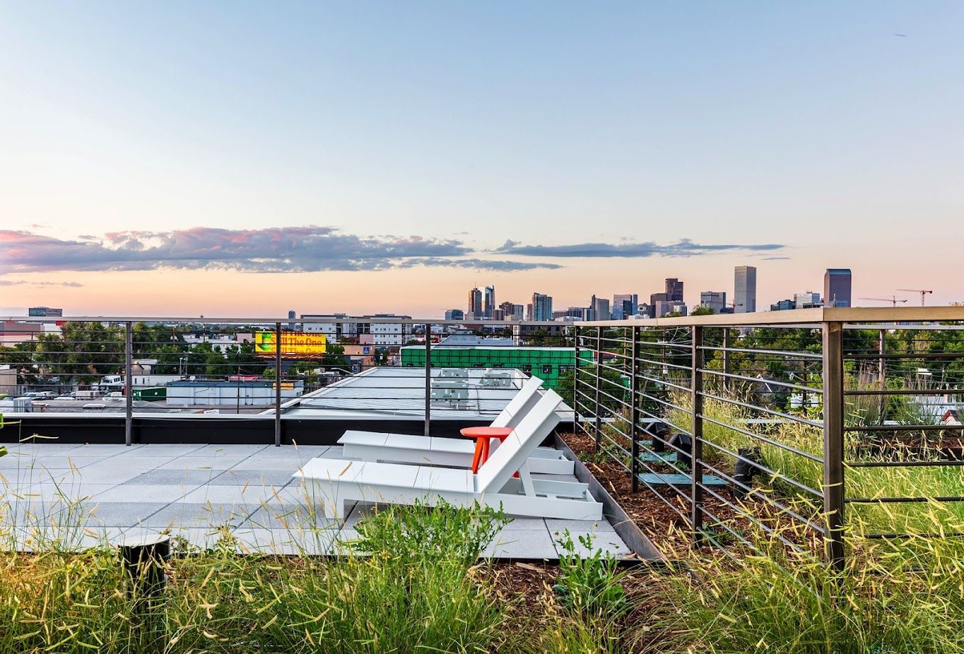 Rooftop view of a city skyline at sunset, featuring a green lawn, white benches, and metal railings.