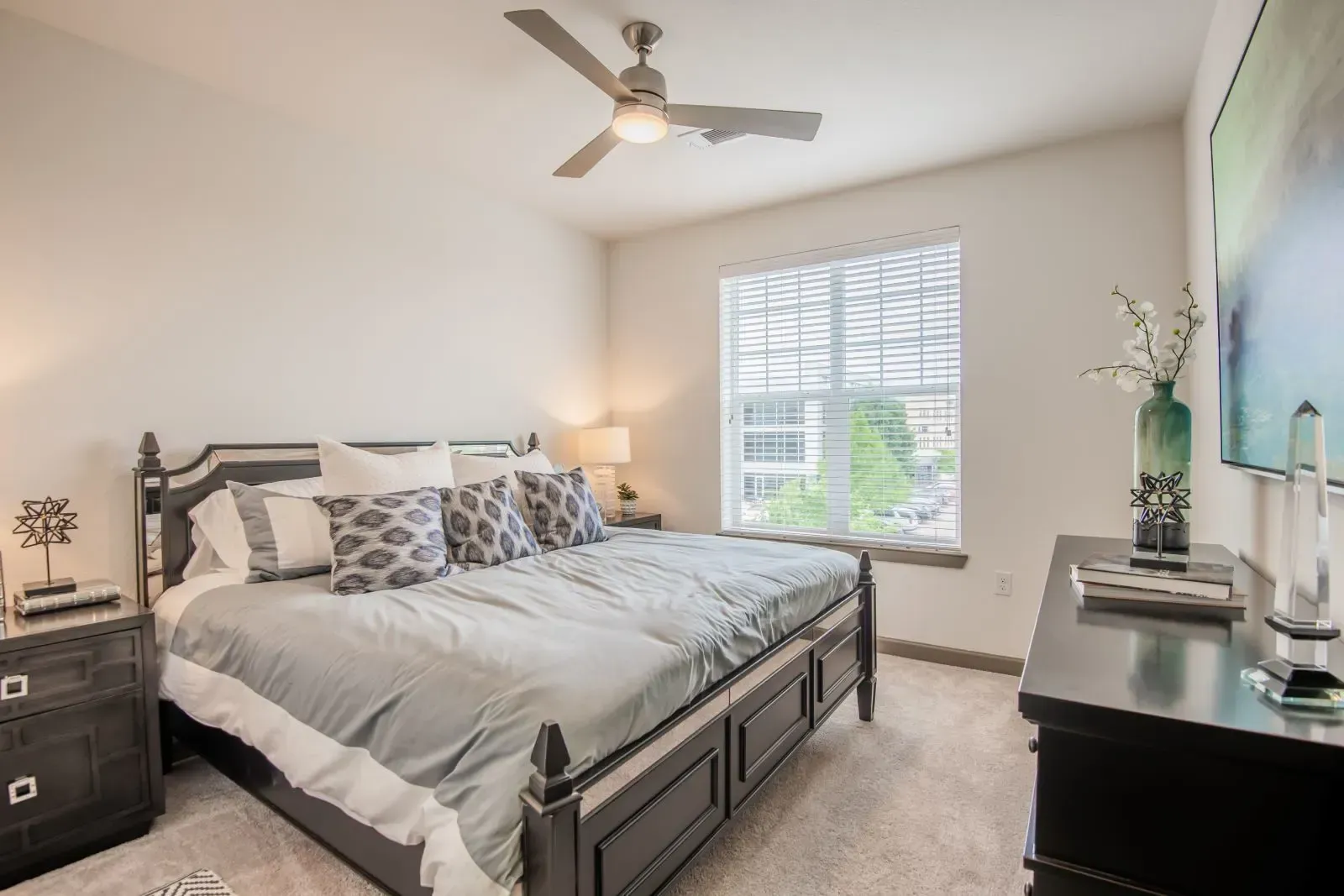 Bedroom in an apartment with a dark wood bed, matching nightstands, ceiling fan, and a large window with blinds.