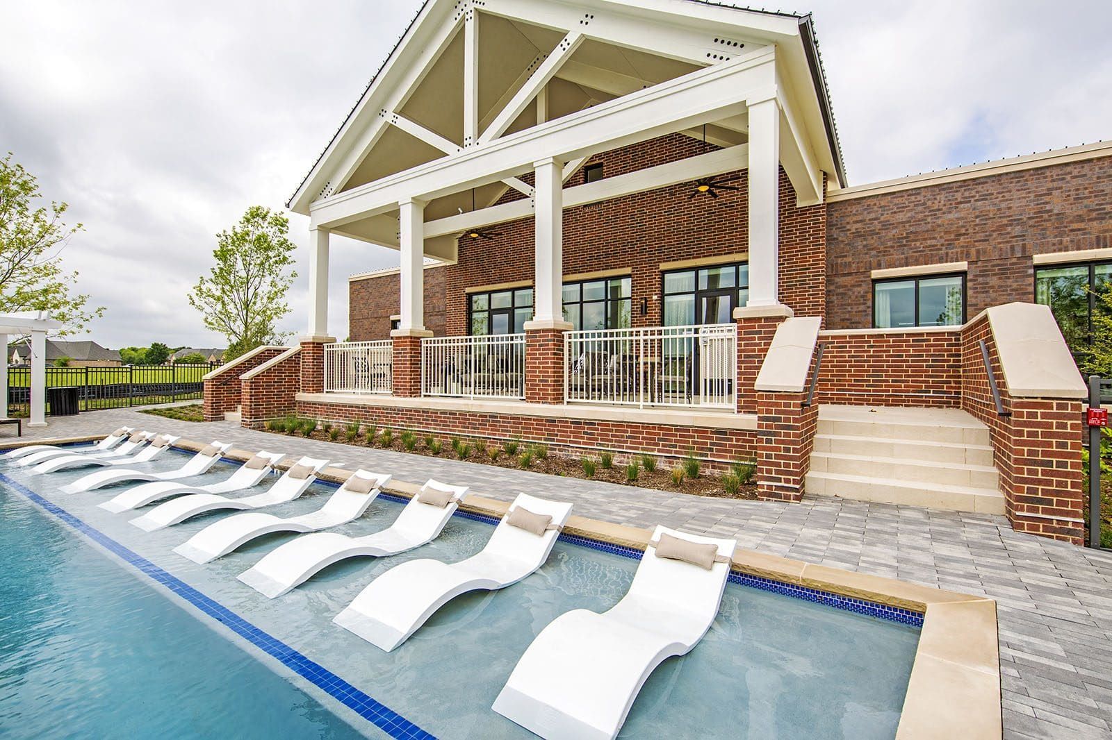 Outdoor pool area with curved white lounge chairs beside a brick clubhouse.