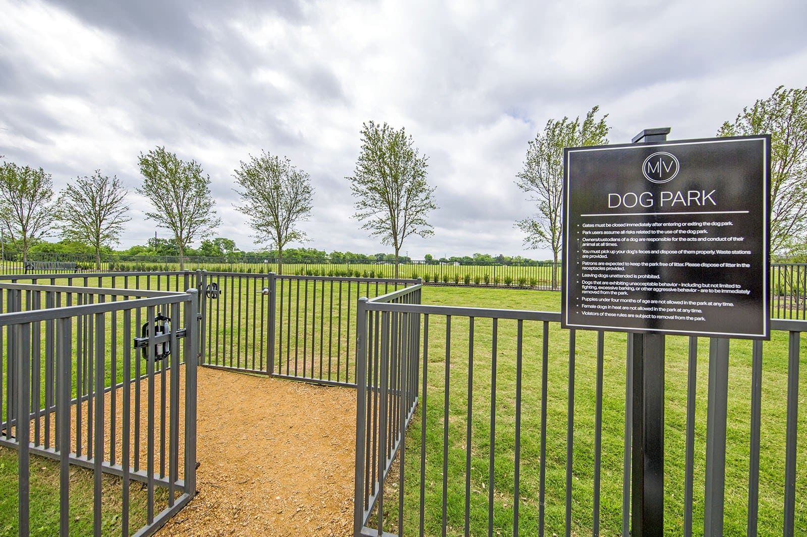 Gated dog park enclosure with gravel path, fence, sign, and open grassy area with trees.