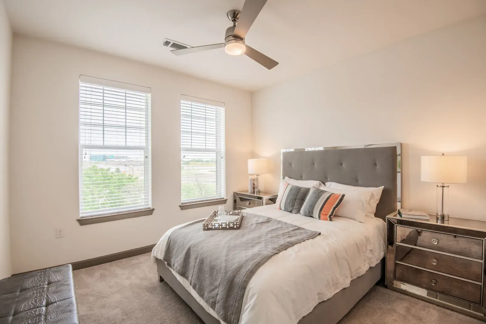 Bedroom with a queen bed, gray tufted headboard, two mirrored nightstands, and large windows with blinds.