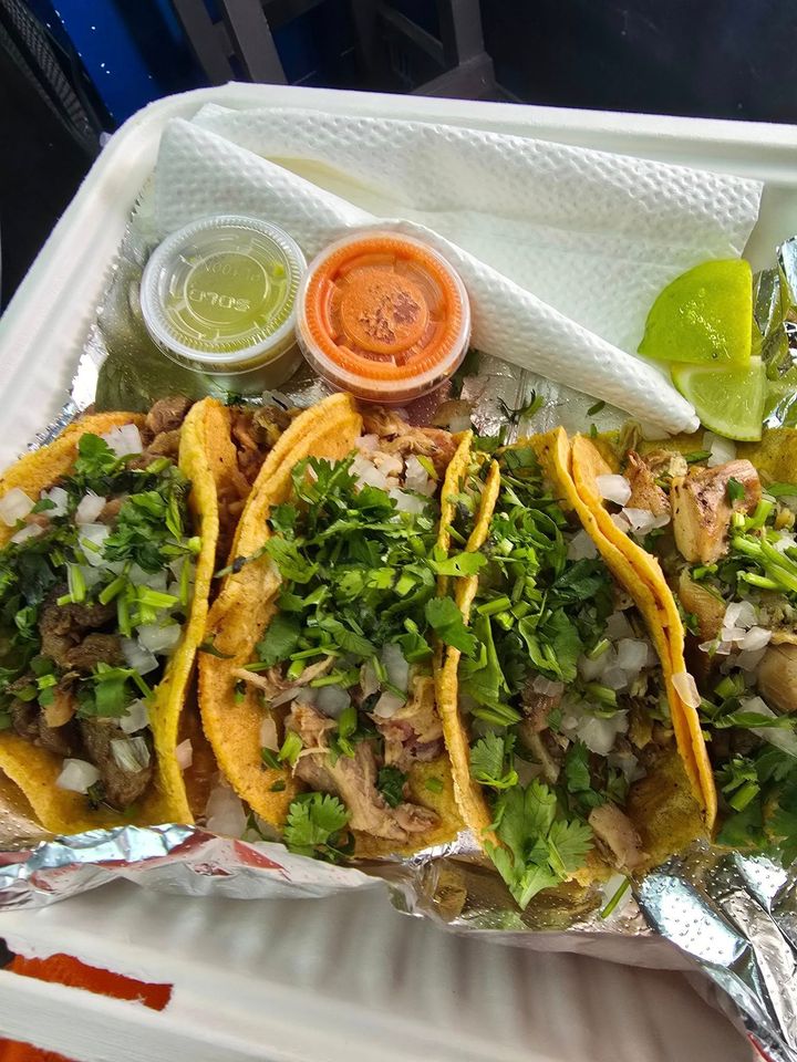 Three street tacos with cilantro and onions in a foil tray, with green salsa and orange salsa cups