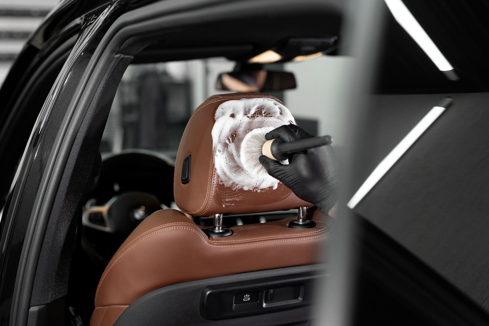 A gloved hand scrubs a leather car headrest with a brush and cleaning foam.