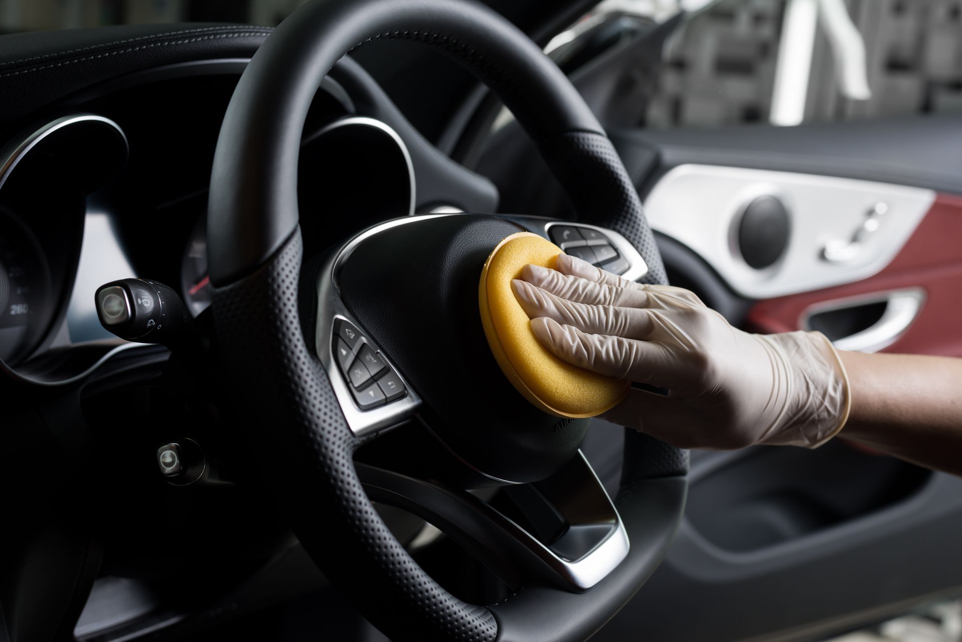 Gloved hand wiping a steering wheel with a yellow sponge inside a car.