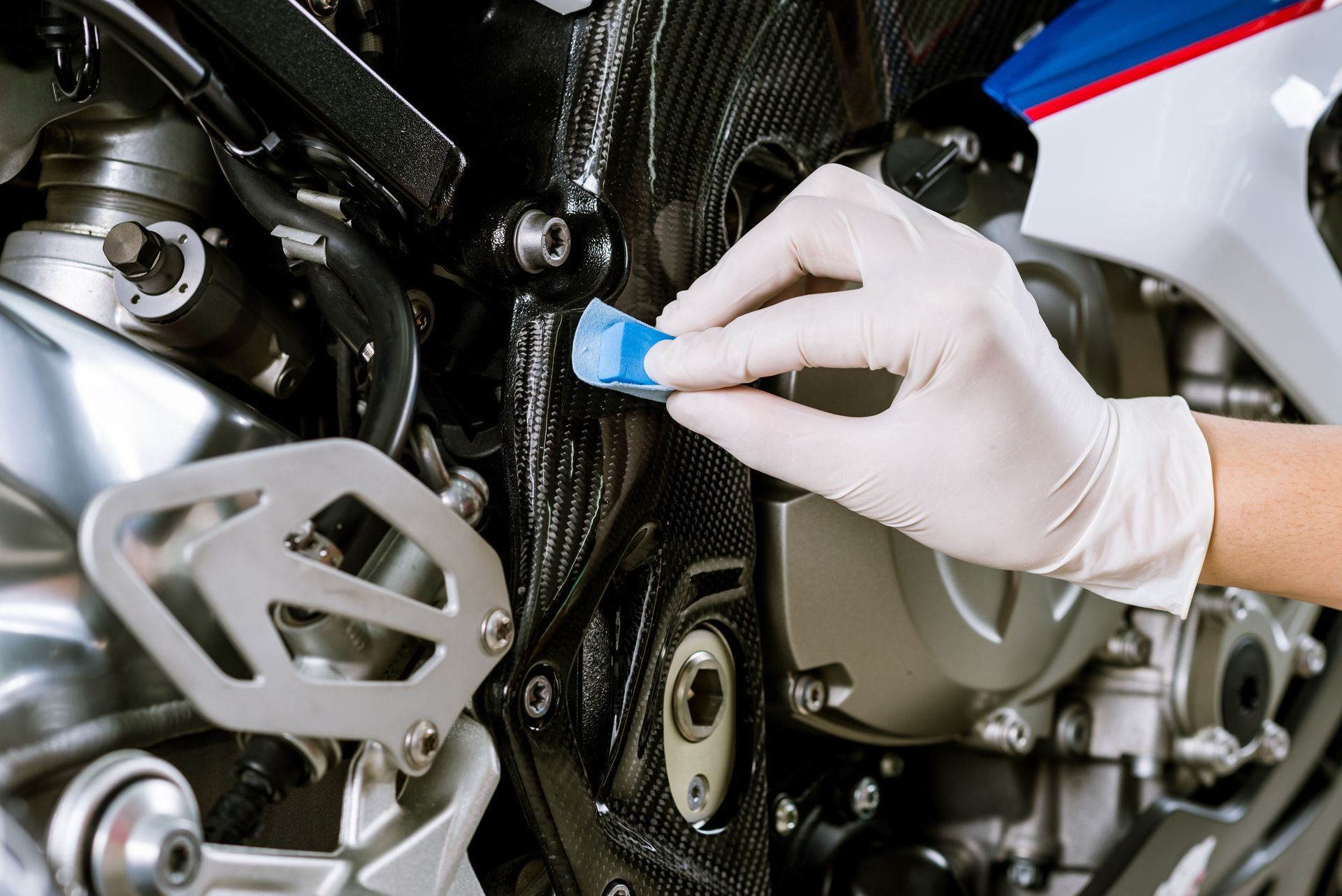 Gloved hand applying a blue product to a black motorcycle frame. Close-up shot with other parts visible.