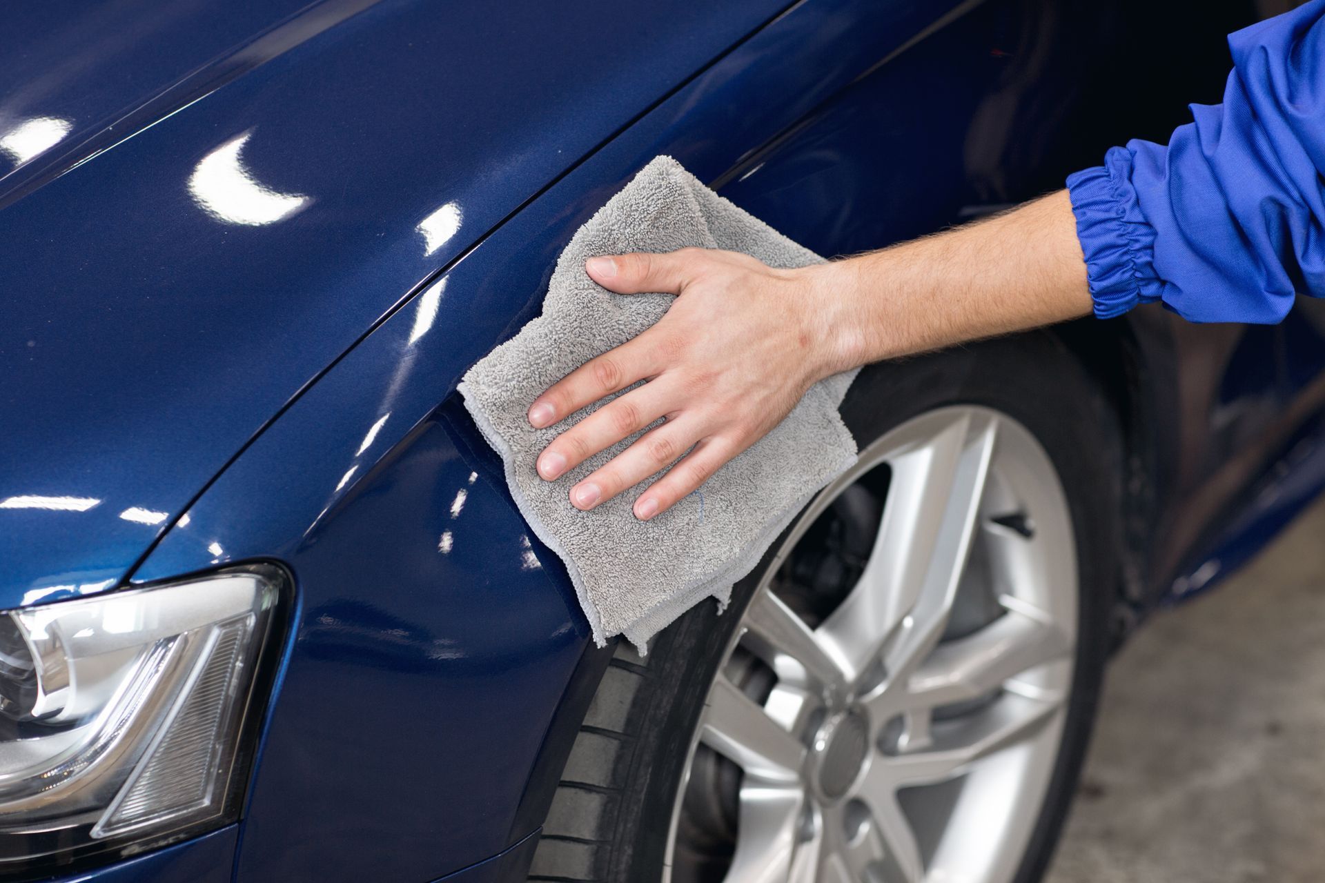 Hand wiping a blue car's hood with a gray microfiber cloth; tire and wheel visible in the background.