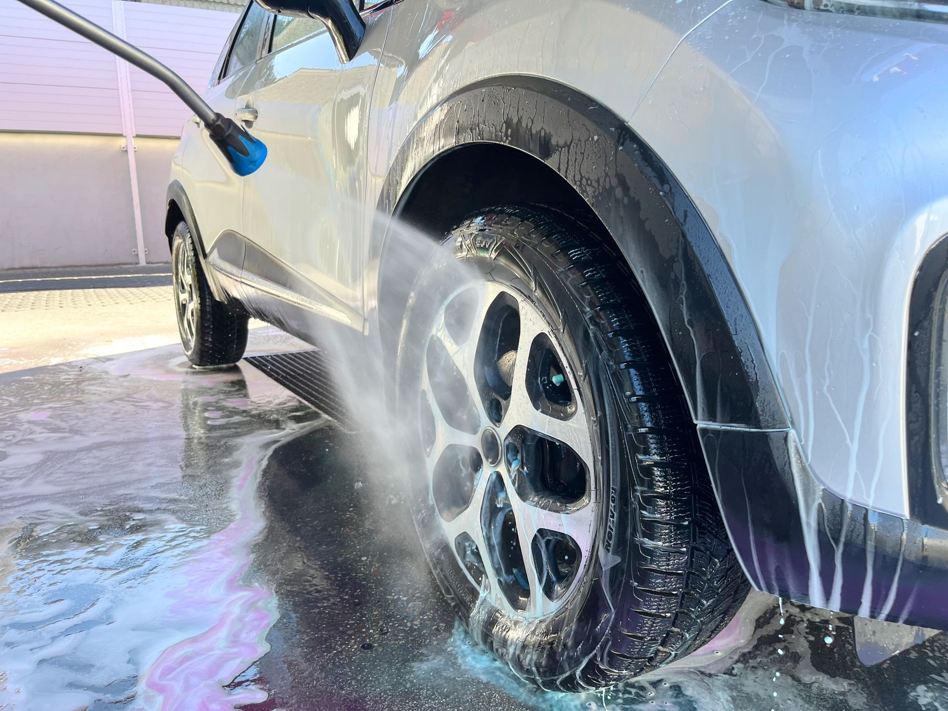 A person washing a white car with a pressure washer; water spraying onto the tire and wheel.