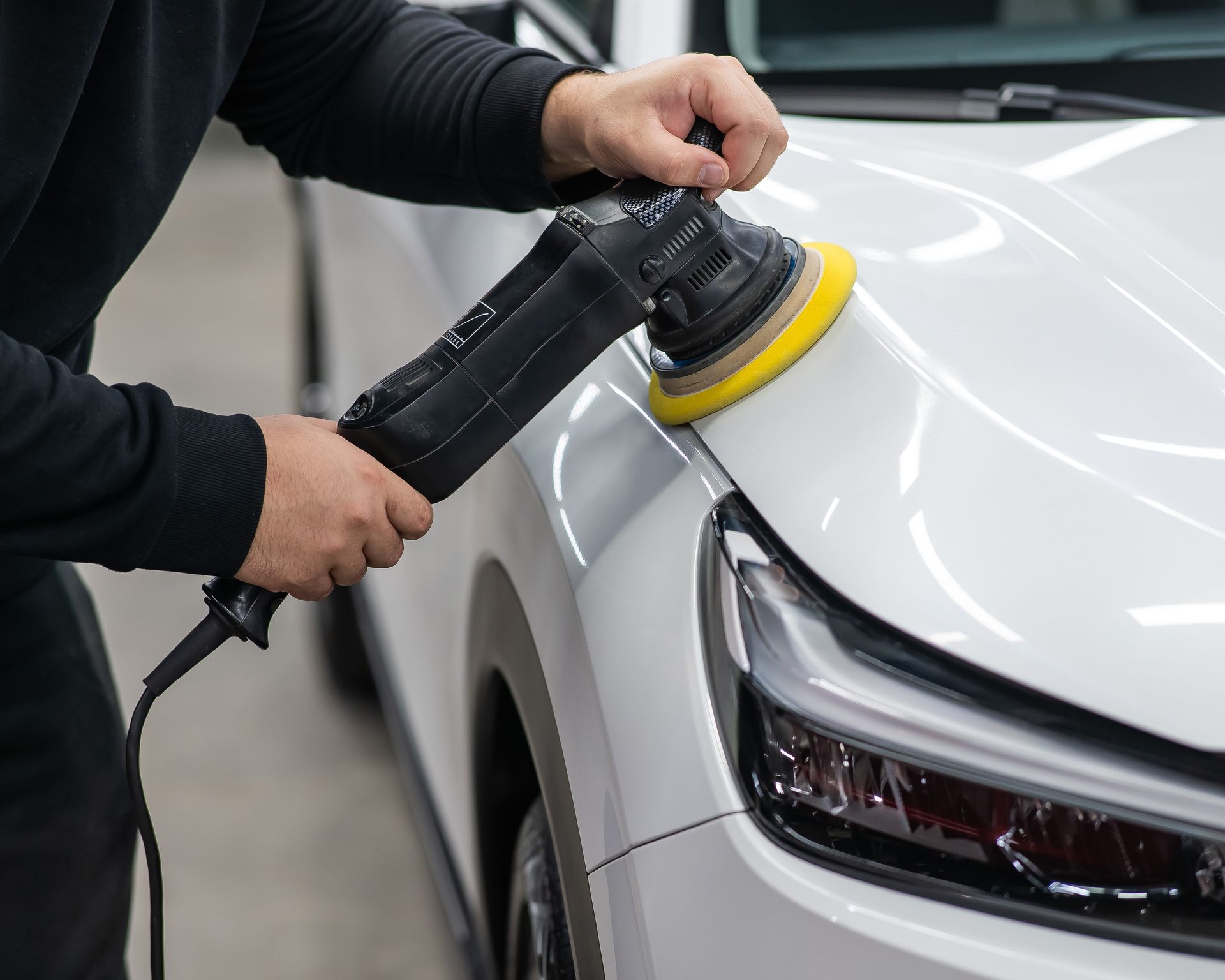 Person polishing white car with an electric buffer in a garage.