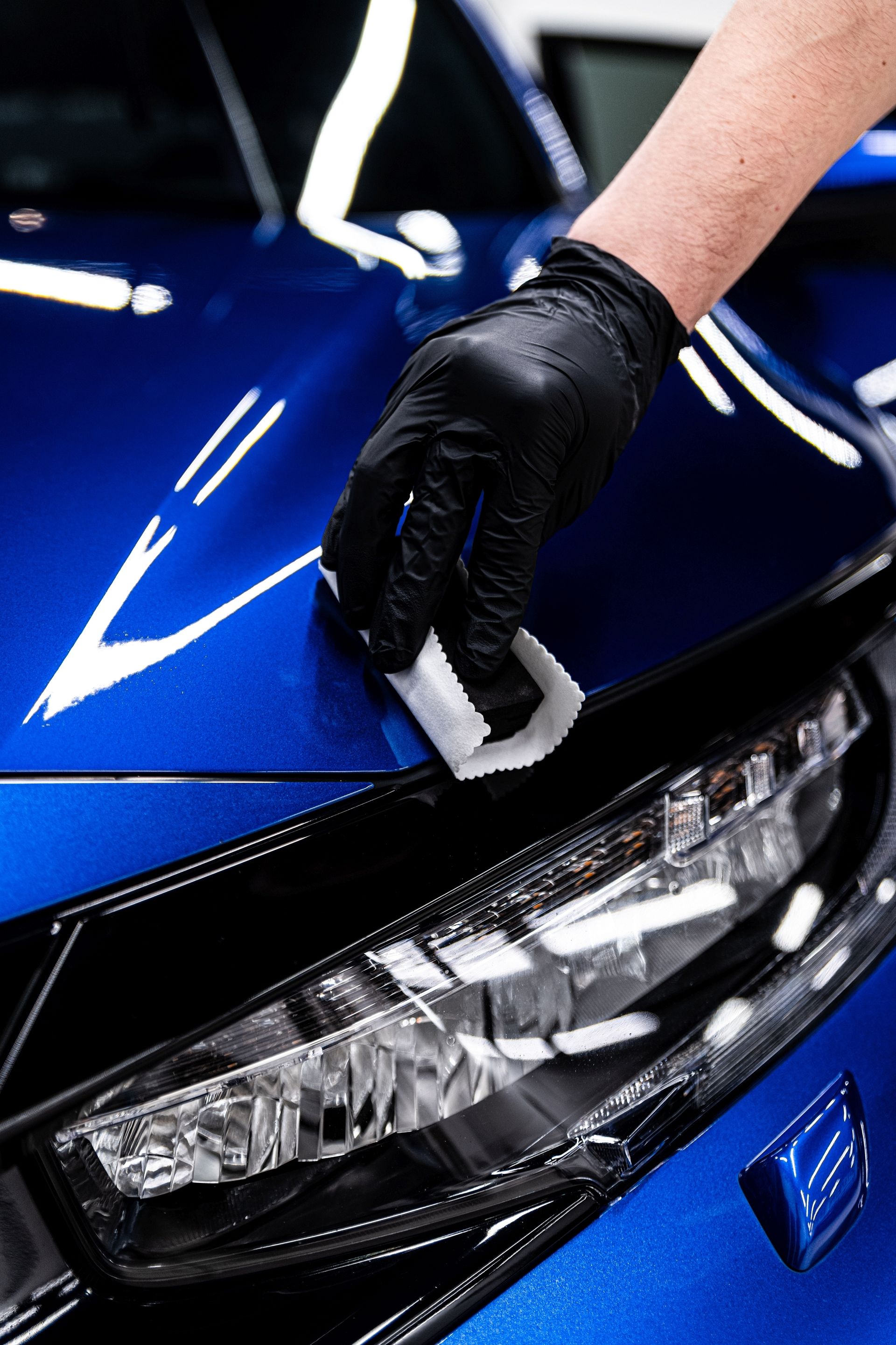 Person wearing a black glove applying car wax to a shiny blue car's hood.