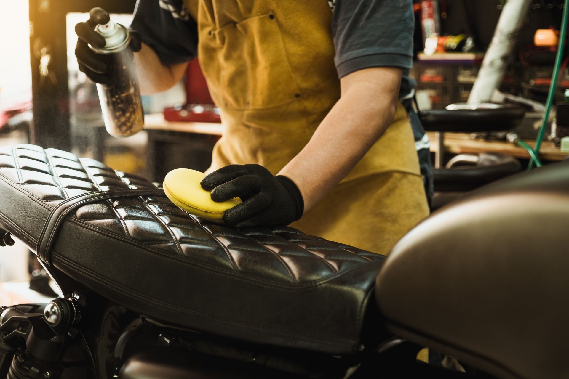 Person cleaning a black motorcycle seat with a spray bottle and sponge in a workshop.