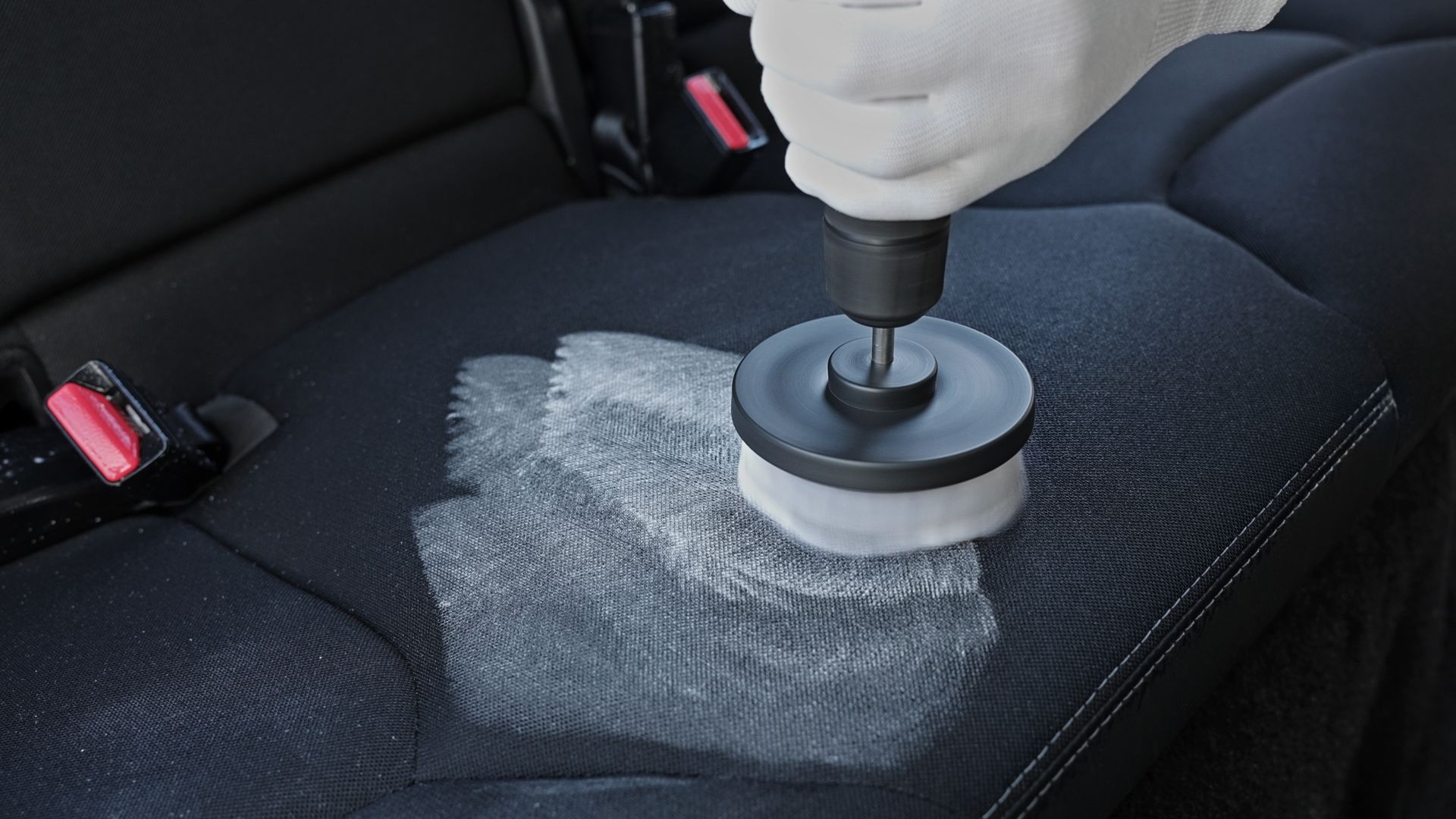 A person uses a drill with a brush attachment to scrub a car seat, covered in cleaning solution.