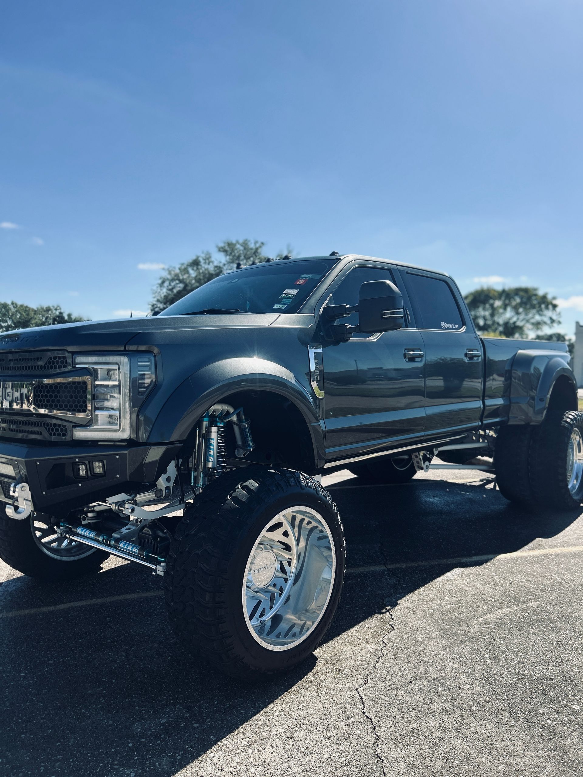 Grey lifted Ford dually truck with chrome wheels parked on pavement under a blue sky.