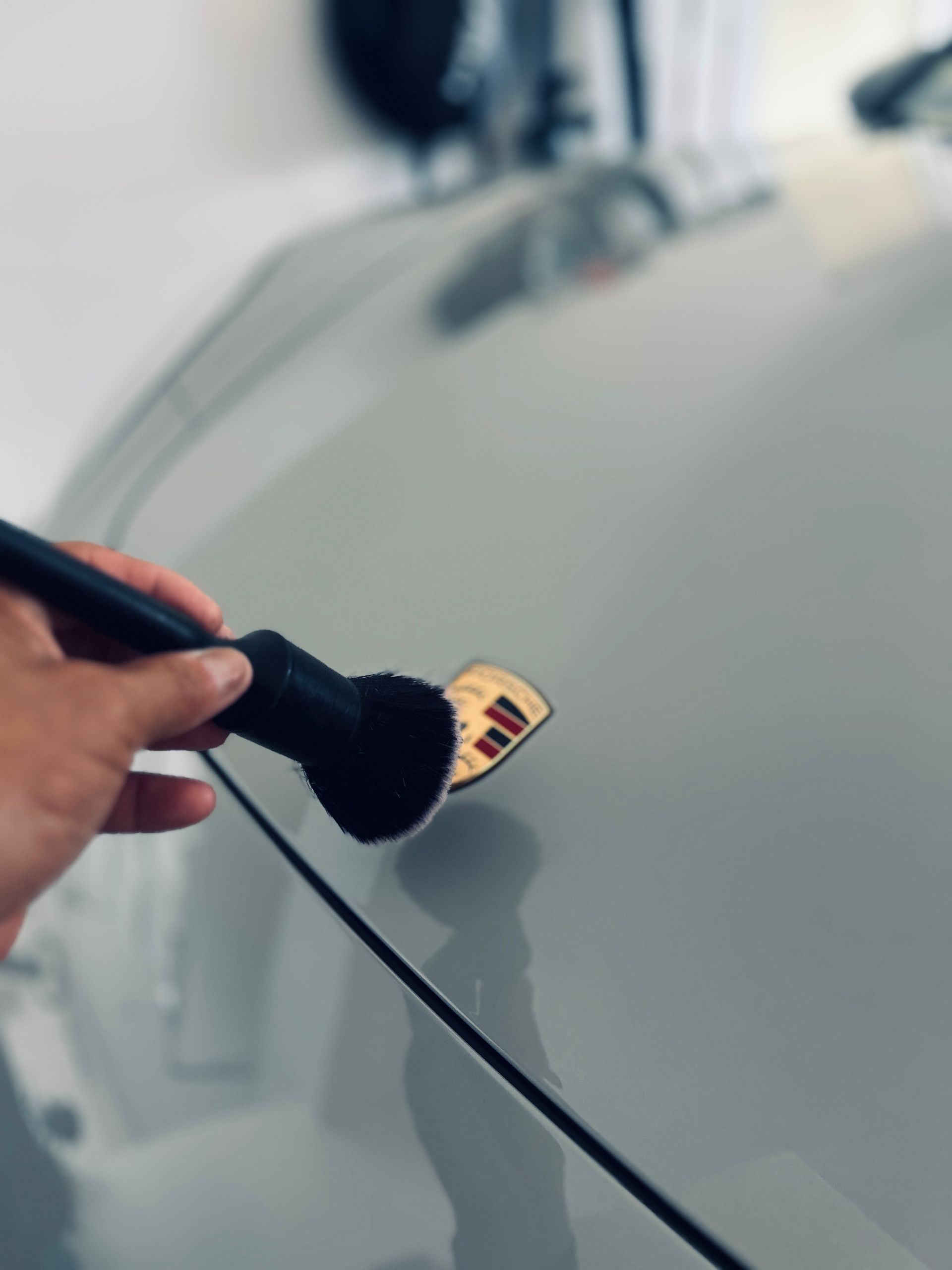 A hand cleaning a Porsche emblem with a black brush on a light gray car.