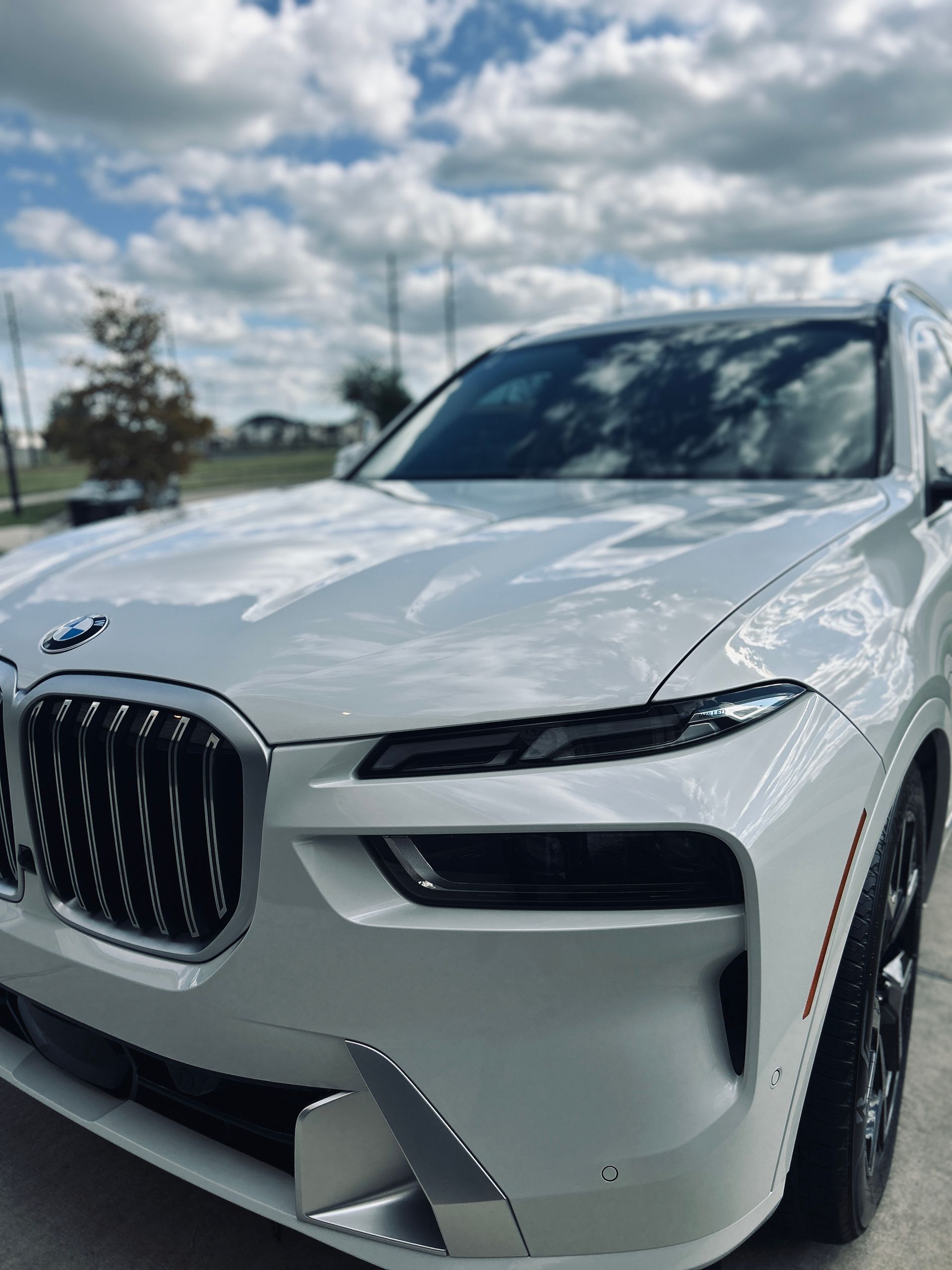 White BMW X7 SUV parked outdoors, shiny finish, against a cloudy sky.