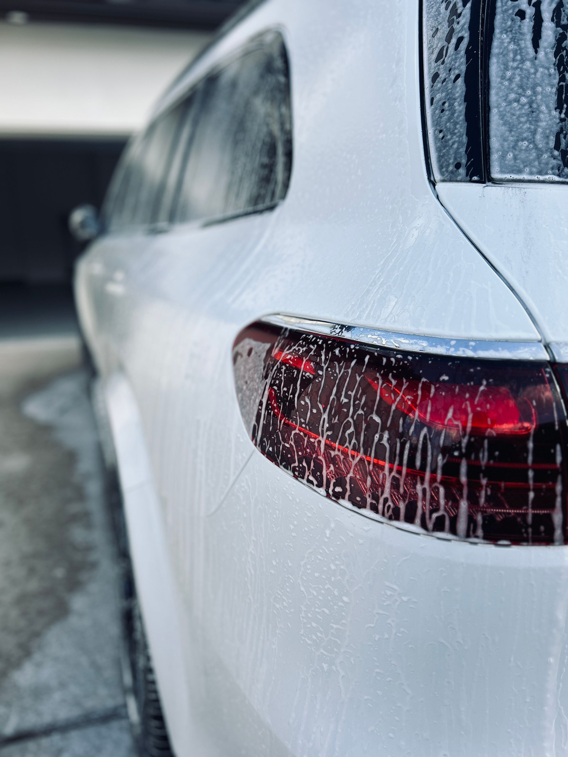 White car's rear quarter panel and tail light covered in soap suds, during washing outdoors.