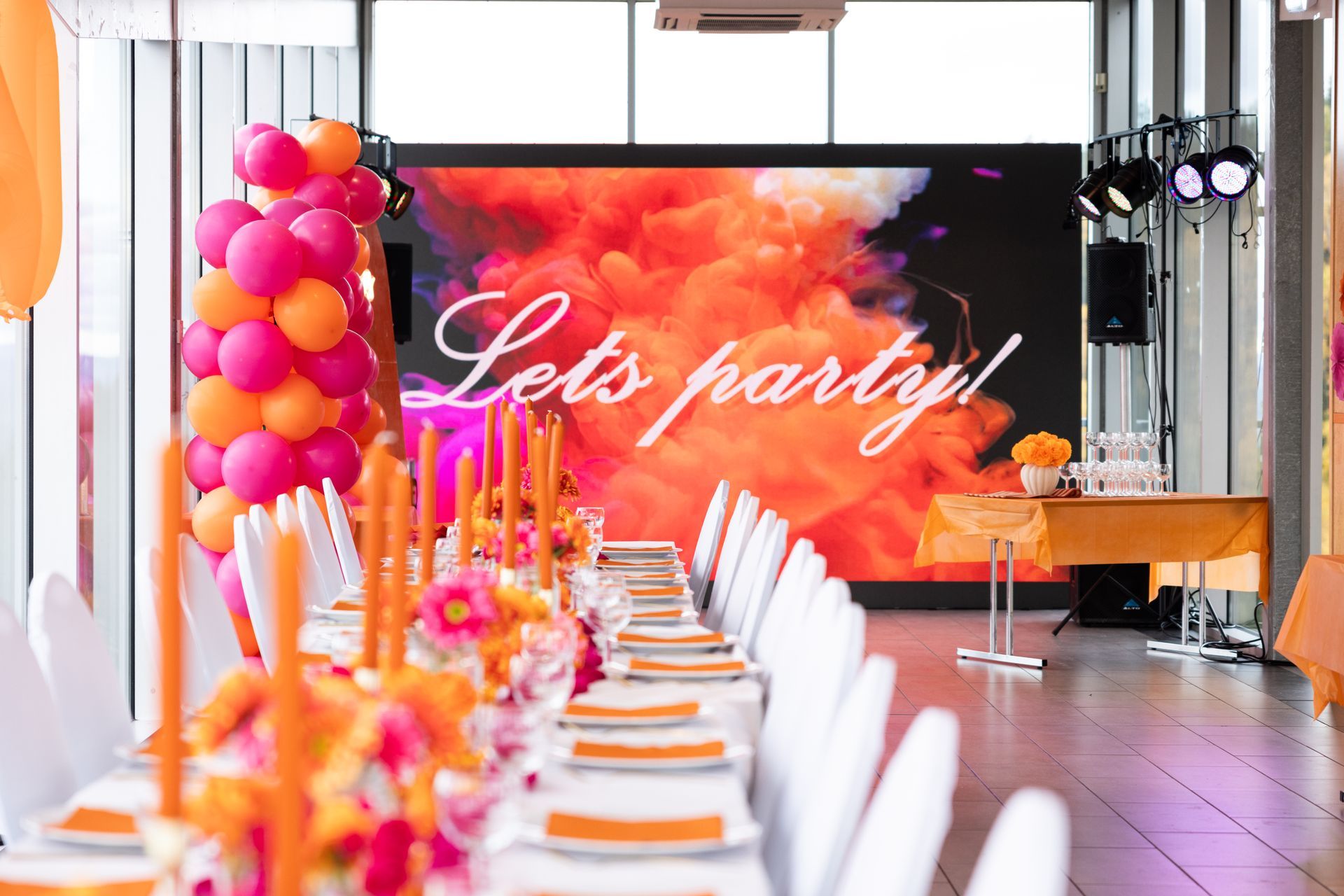 Festoppsett med langt bord, fargerik dekorasjon og en skjerm hvor det står «Lets fest!».