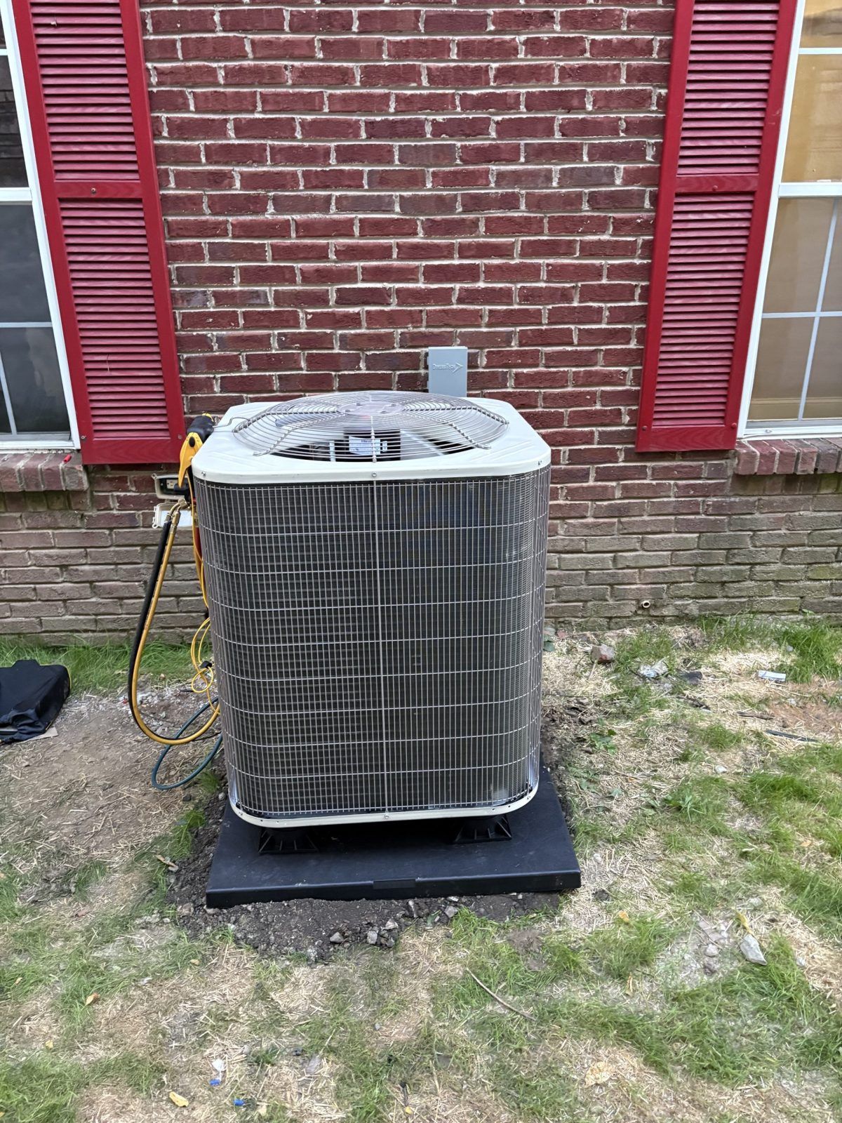 Air conditioning unit outside a brick building, red shutters, resting on a black pad.
