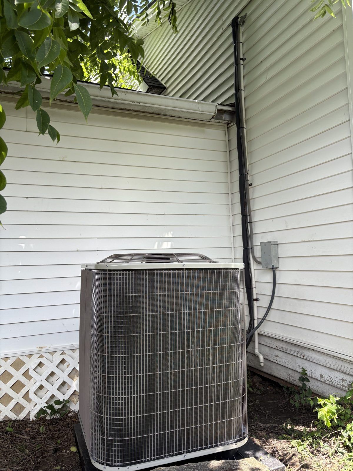 AC unit outside a white house. Black pipes and gray electrical box are attached to the wall.