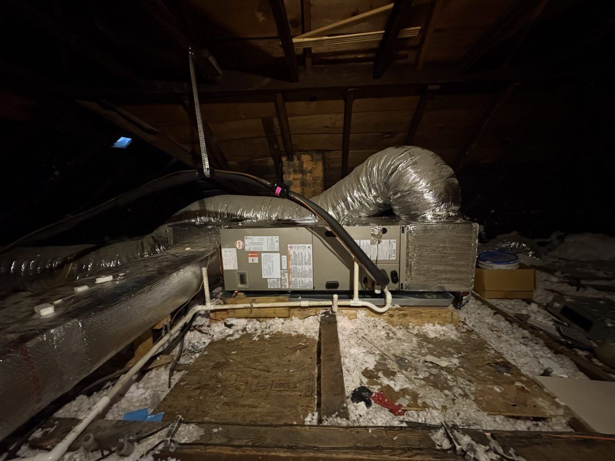 HVAC unit in an attic. Metallic ductwork, insulation, and exposed rafters. Dark setting, poor lighting.