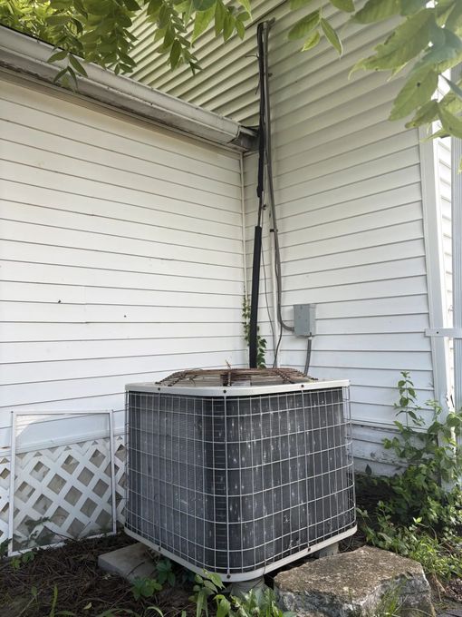 Outdoor air conditioning unit against a white house. Black pipes run up the siding.