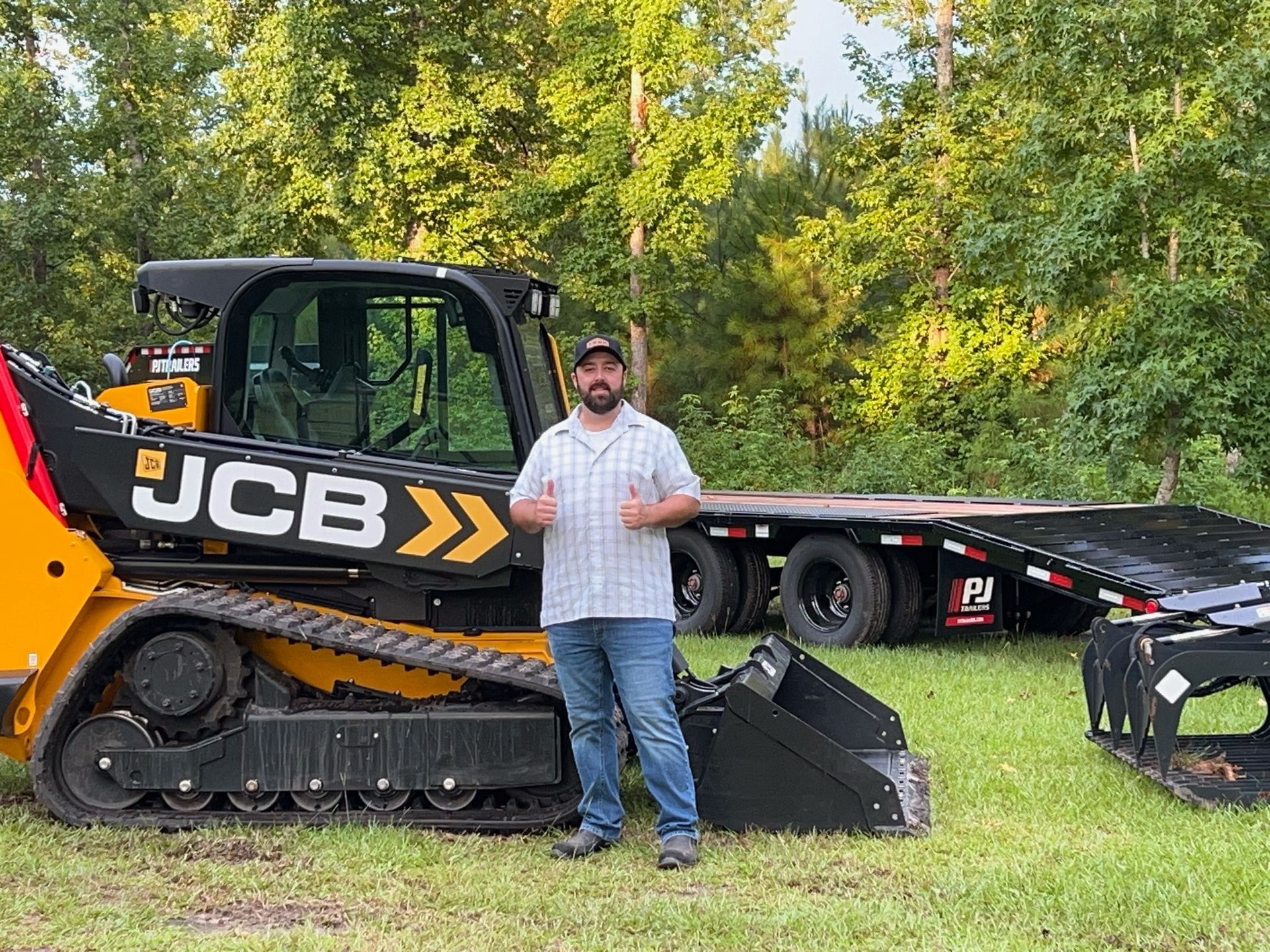 Man giving thumbs up next to yellow JCB track loader and black trailer in grassy area.