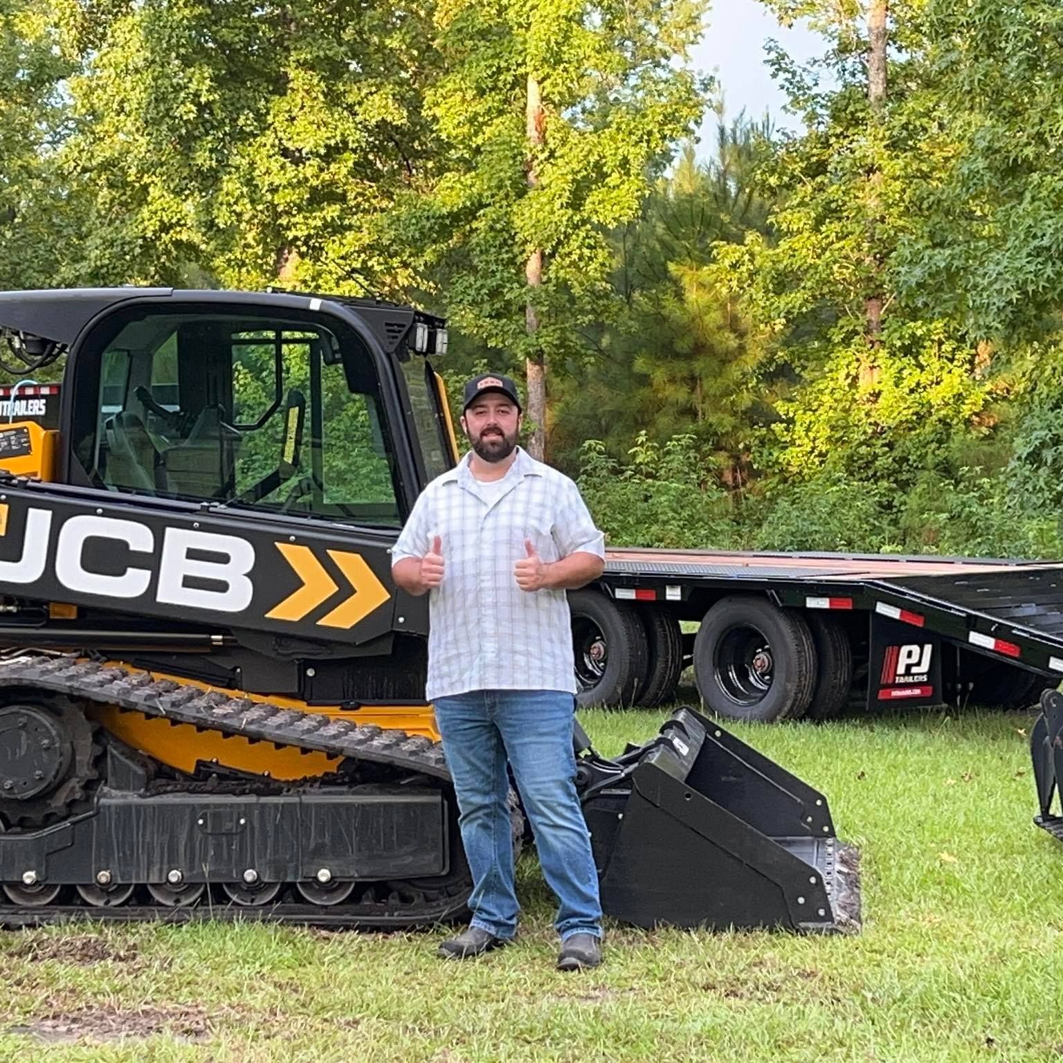 Man giving thumbs up stands next to a yellow JCB track skid steer and flatbed trailer outdoors.