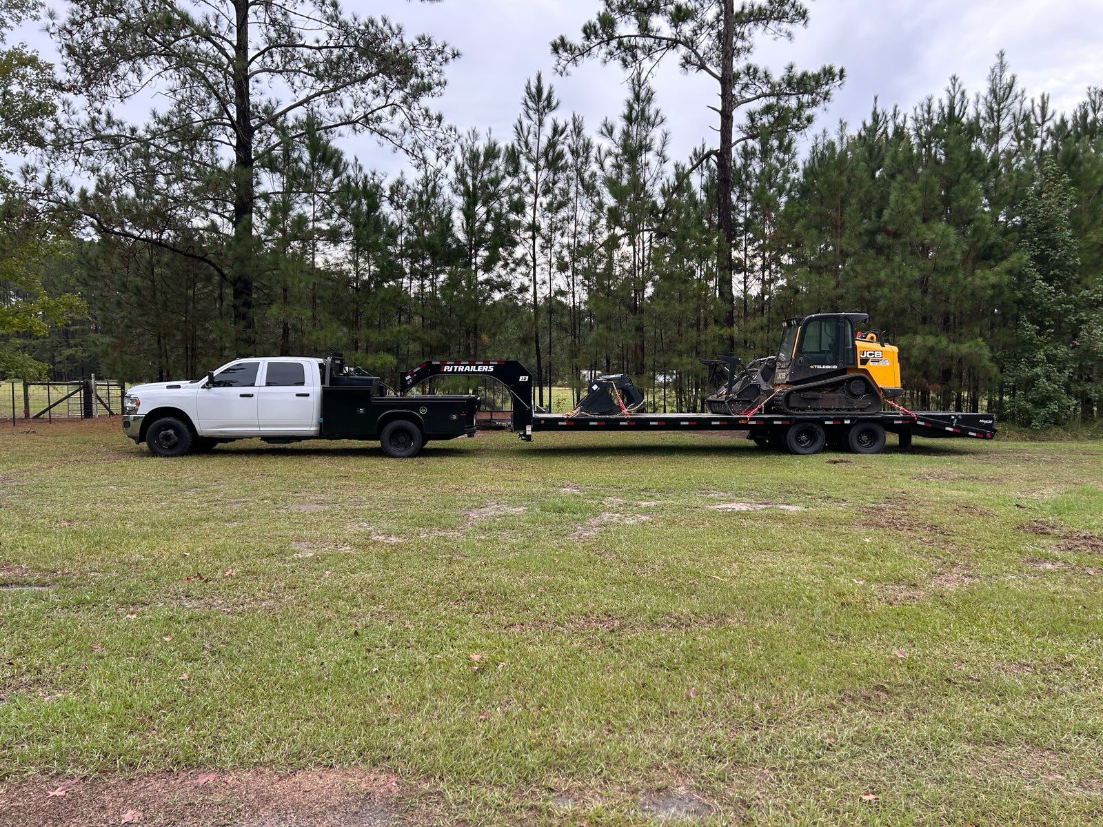 White truck towing a flatbed trailer with heavy machinery on a grassy field near trees.