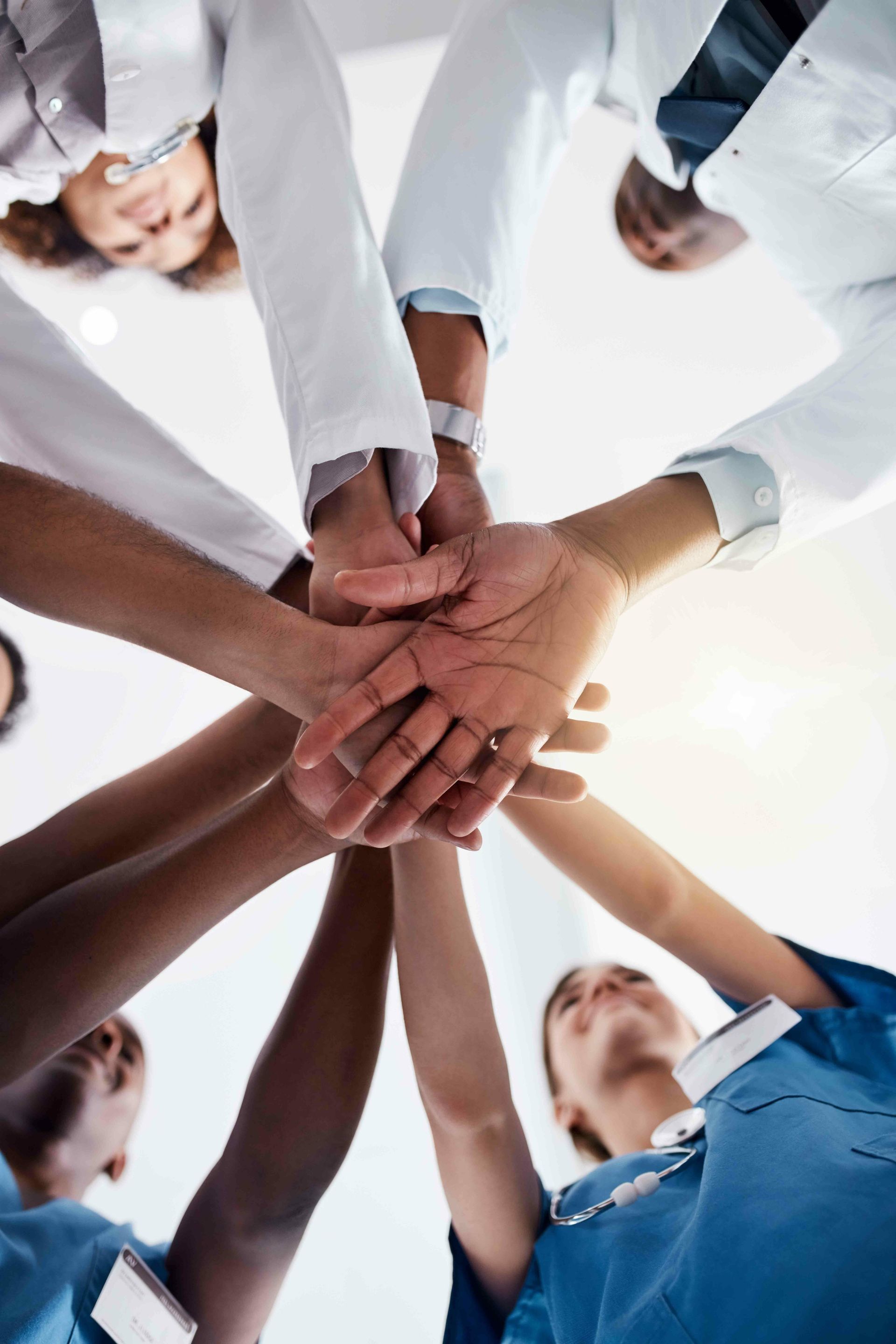 Doctors and nurses in various scrubs and lab coats put hands together in a team huddle.