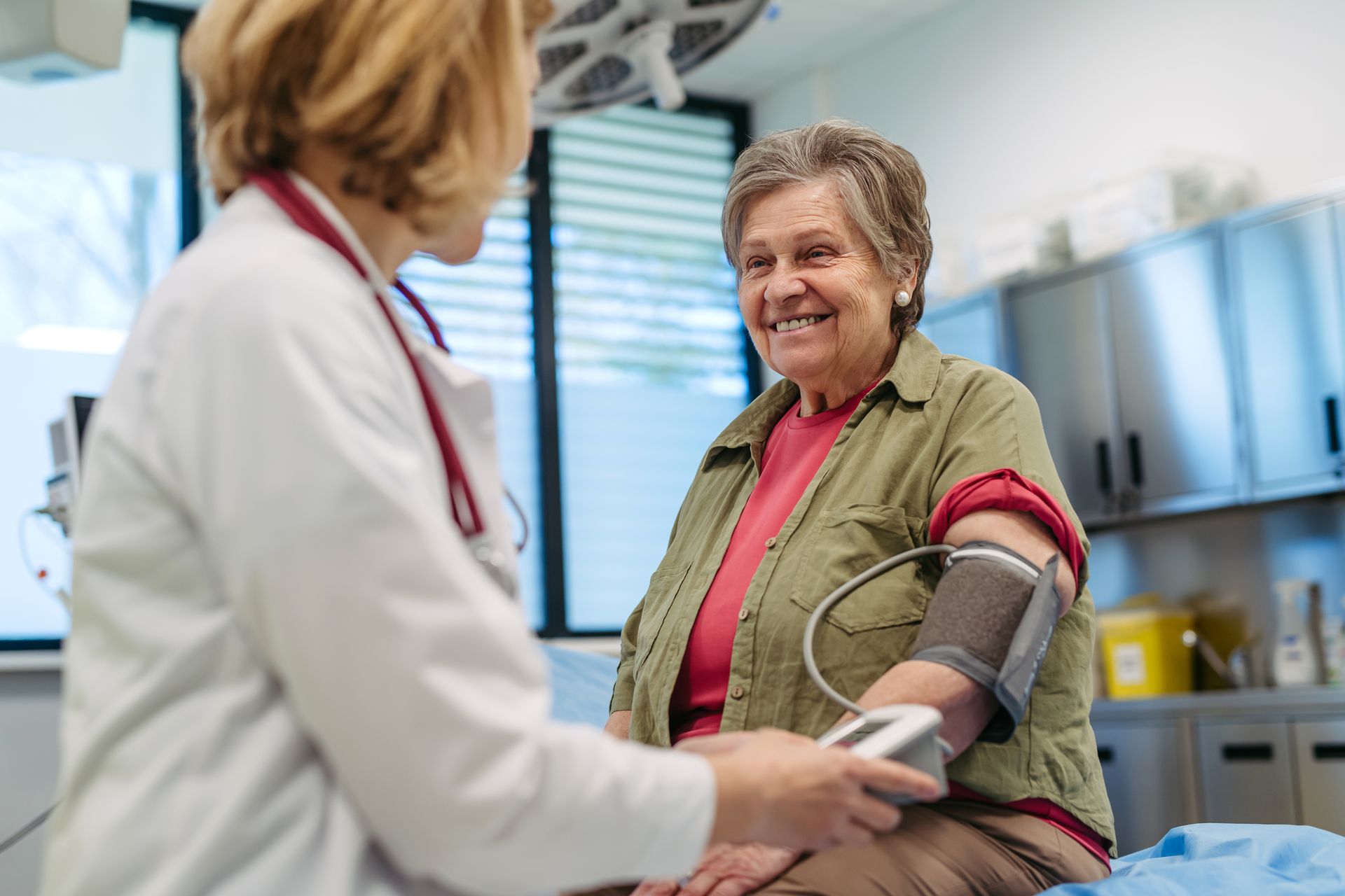 Doctor taking an elderly woman's blood pressure in a medical office. The woman smiles.