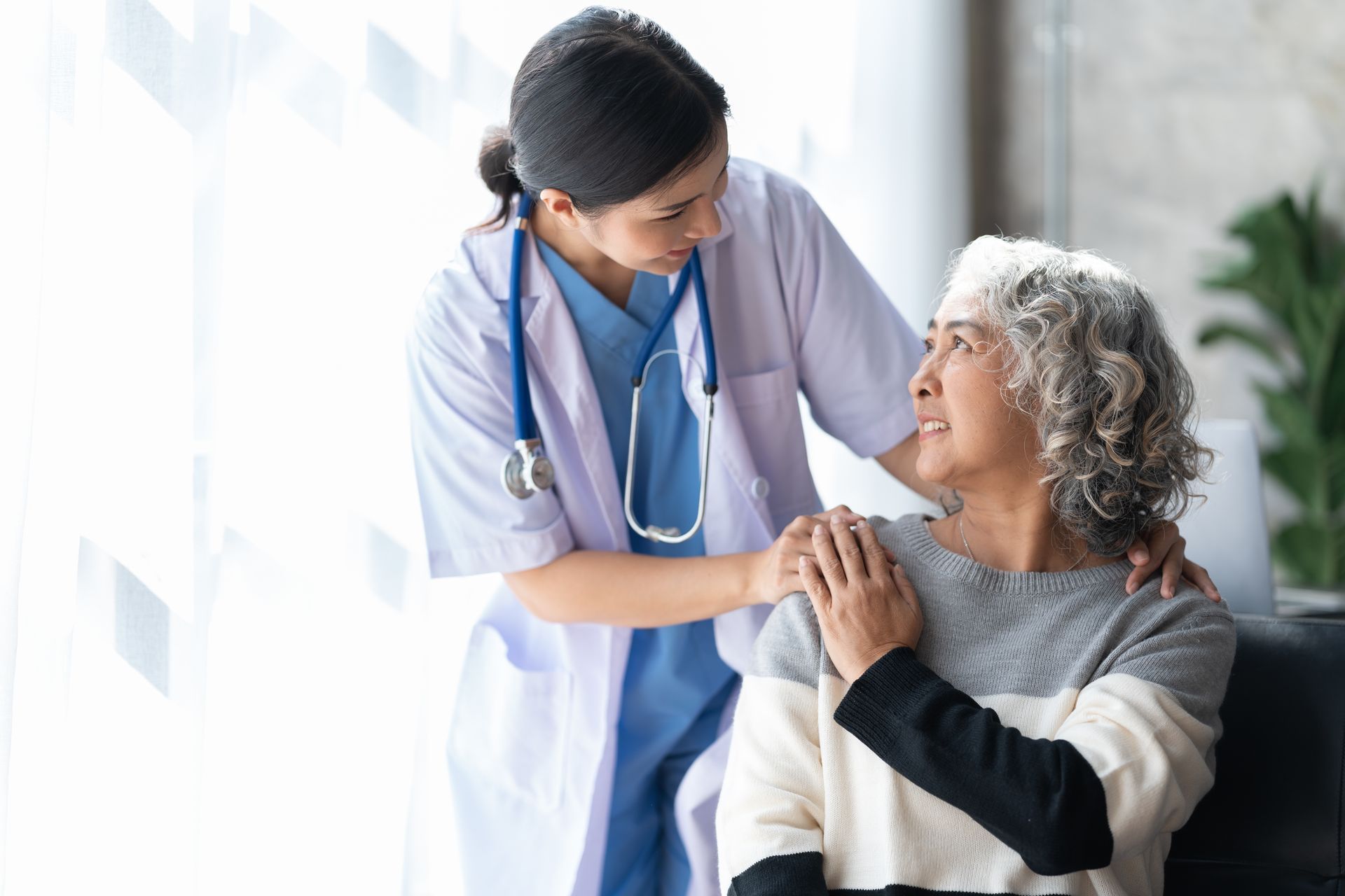 A young female doctor in a white coat comforts an elderly Asian woman with a hand on her shoulder.