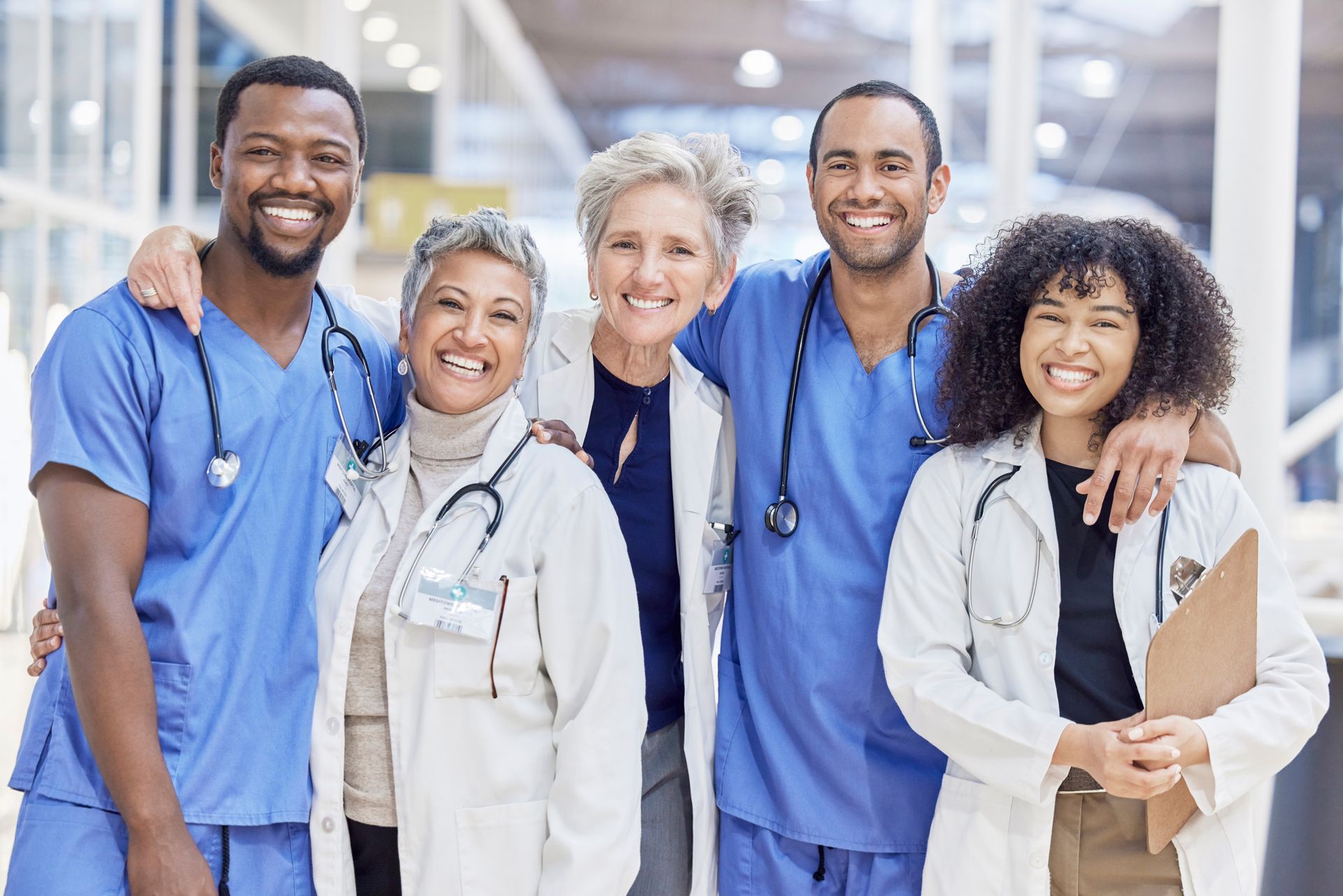 Smiling medical team in blue scrubs and white coats posing indoors, arms around each other.
