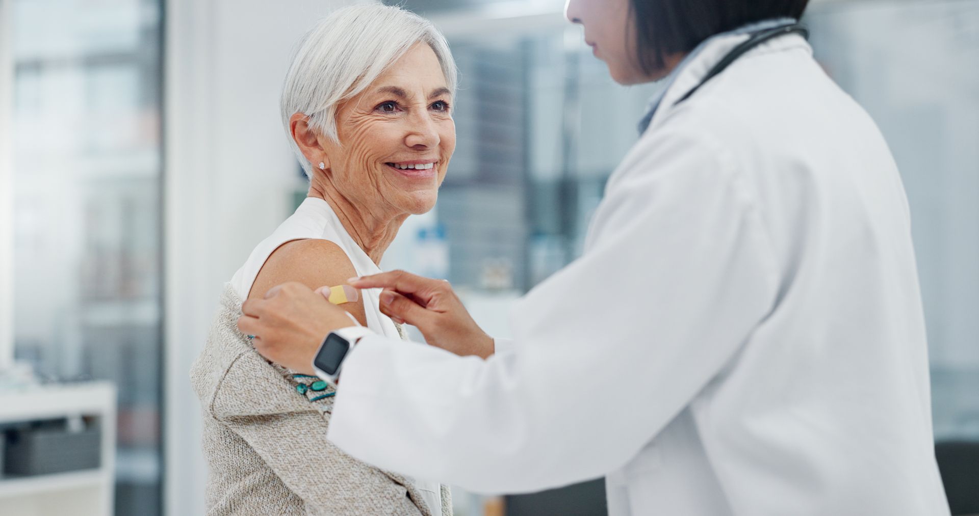 Doctor places bandage on the arm of an older woman after a vaccination; medical office setting.