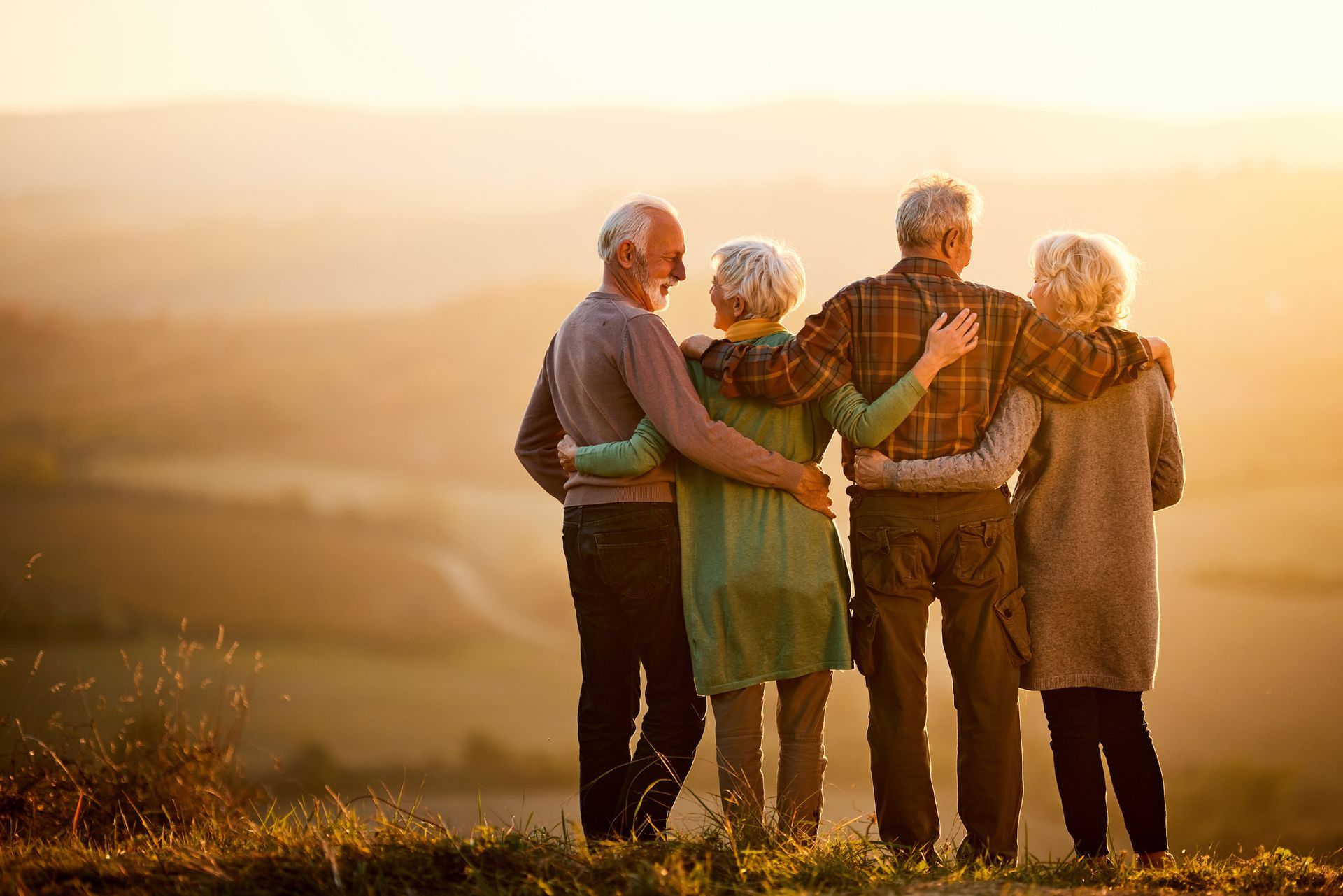 Four older adults with arms around each other, overlooking a golden landscape at sunset.