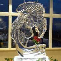 Clear ice sculpture of a man's head kissing a woman's forehead with an inserted red rose in the base indoors against a window backdrop