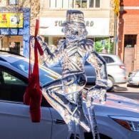 Transparent dancer ice sculpture beside a parked car on a city street, with red toe shoes draped and hanging down from the upper hand and the lower hand holds a cane to the ground.