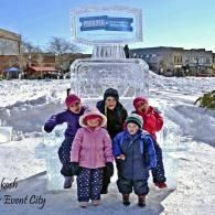 Five children posing in front of a large ice sculpture in a snowy town square
