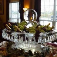 Crystal fruit display on a dining table with grapes and decorative glassware bottles with an ice sculpture in the center by a window